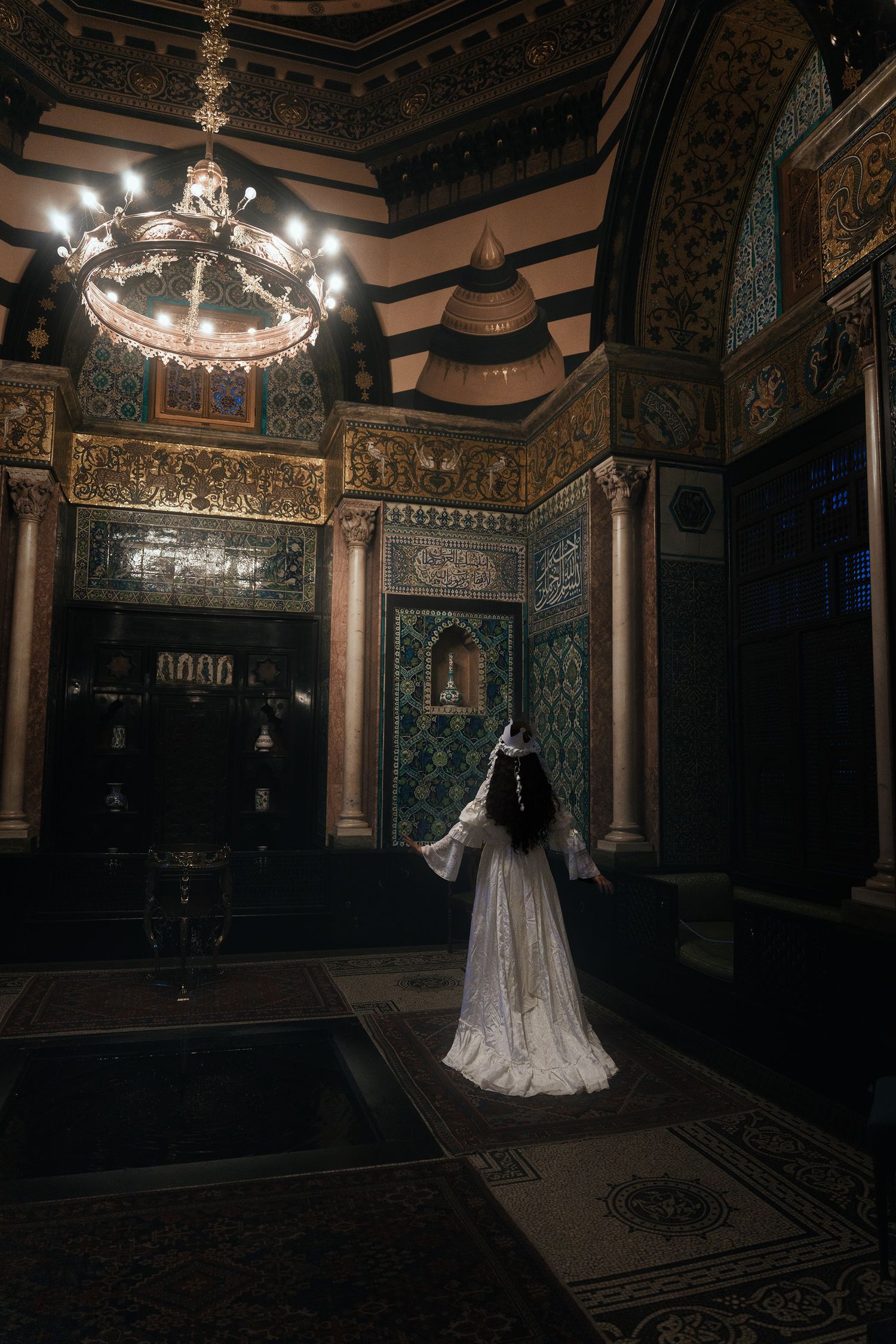Lone figure in white dress gazes up at ornate Islamic-style interior featuring intricate tilework, chandelier, and golden details.
