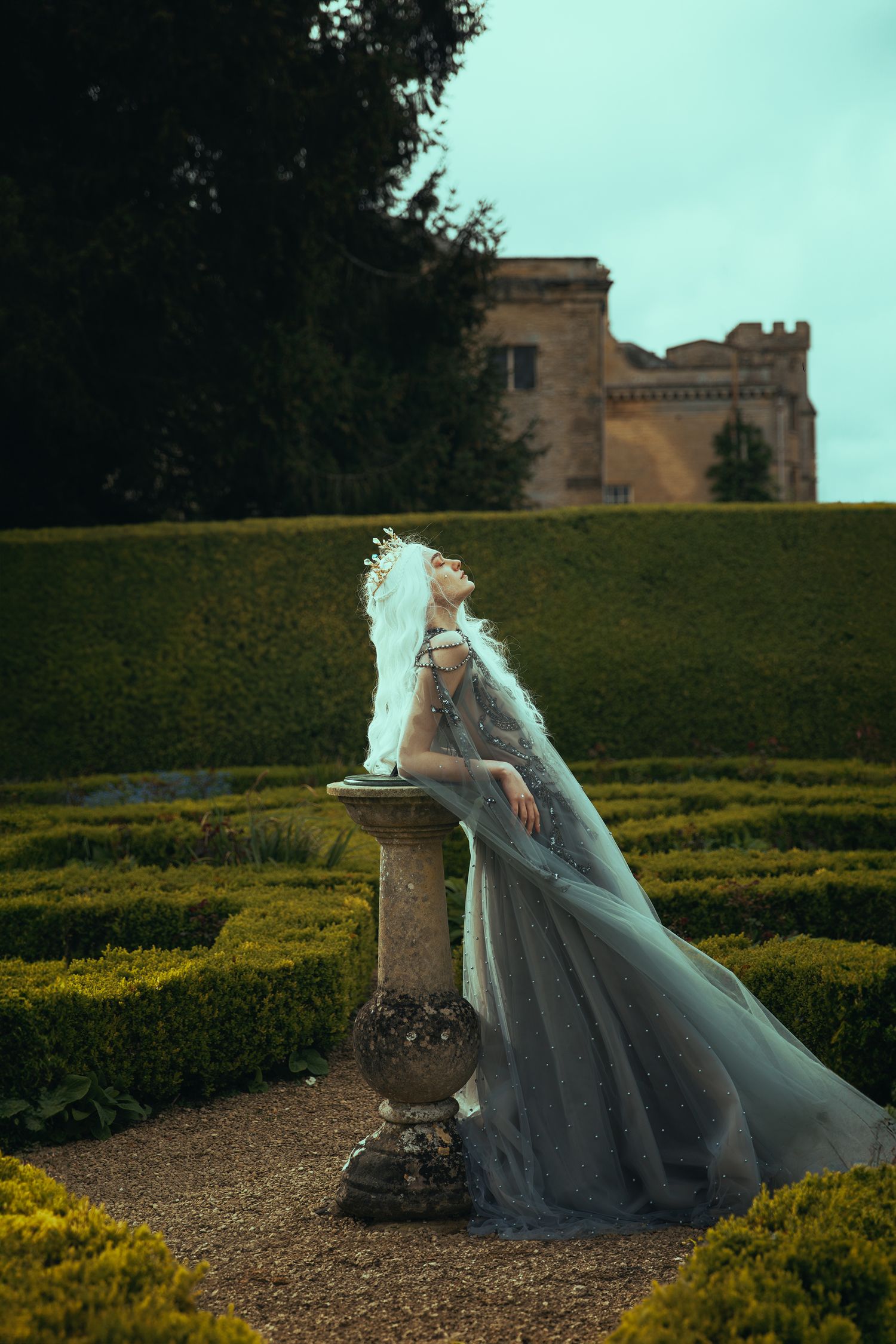 Blonde woman in grey tulle gown and white feather crown poses by stone sundial in castle garden.