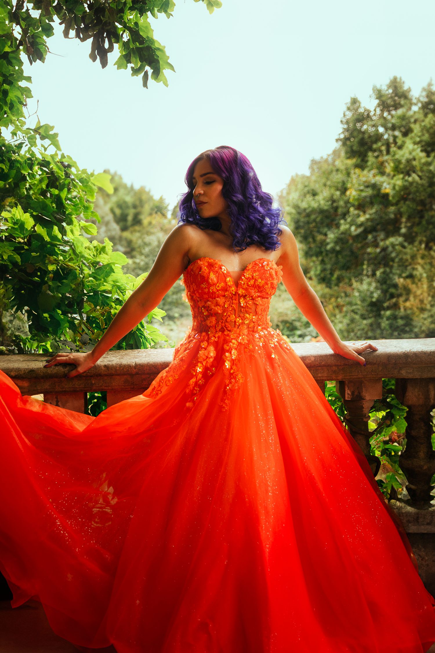 A woman with purple hair poses in a vibrant orange floral ball gown on a balcony surrounded by green trees.