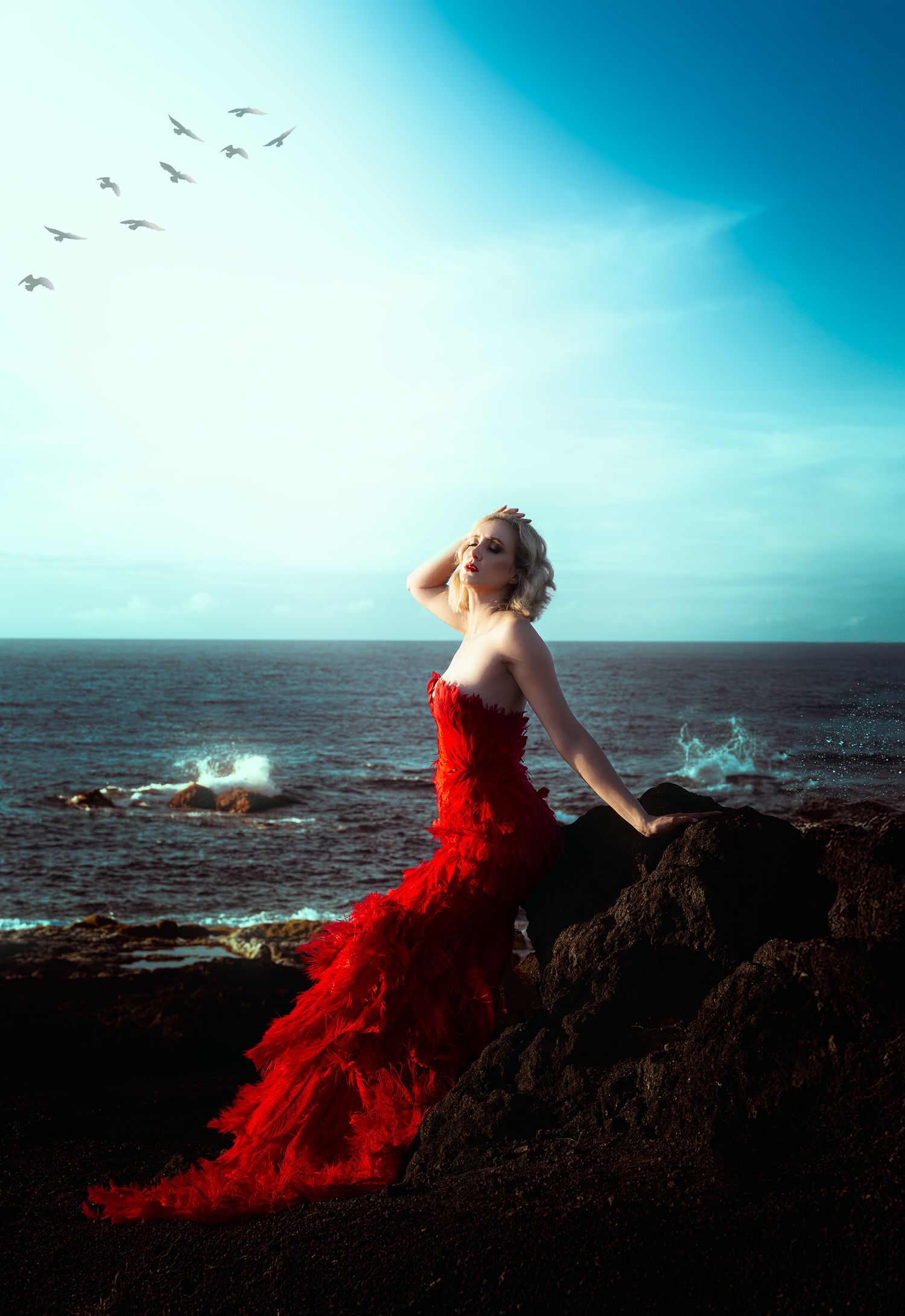 A blonde woman in a flowing red gown sits on ocean rocks gazing upward under a vivid blue sky.