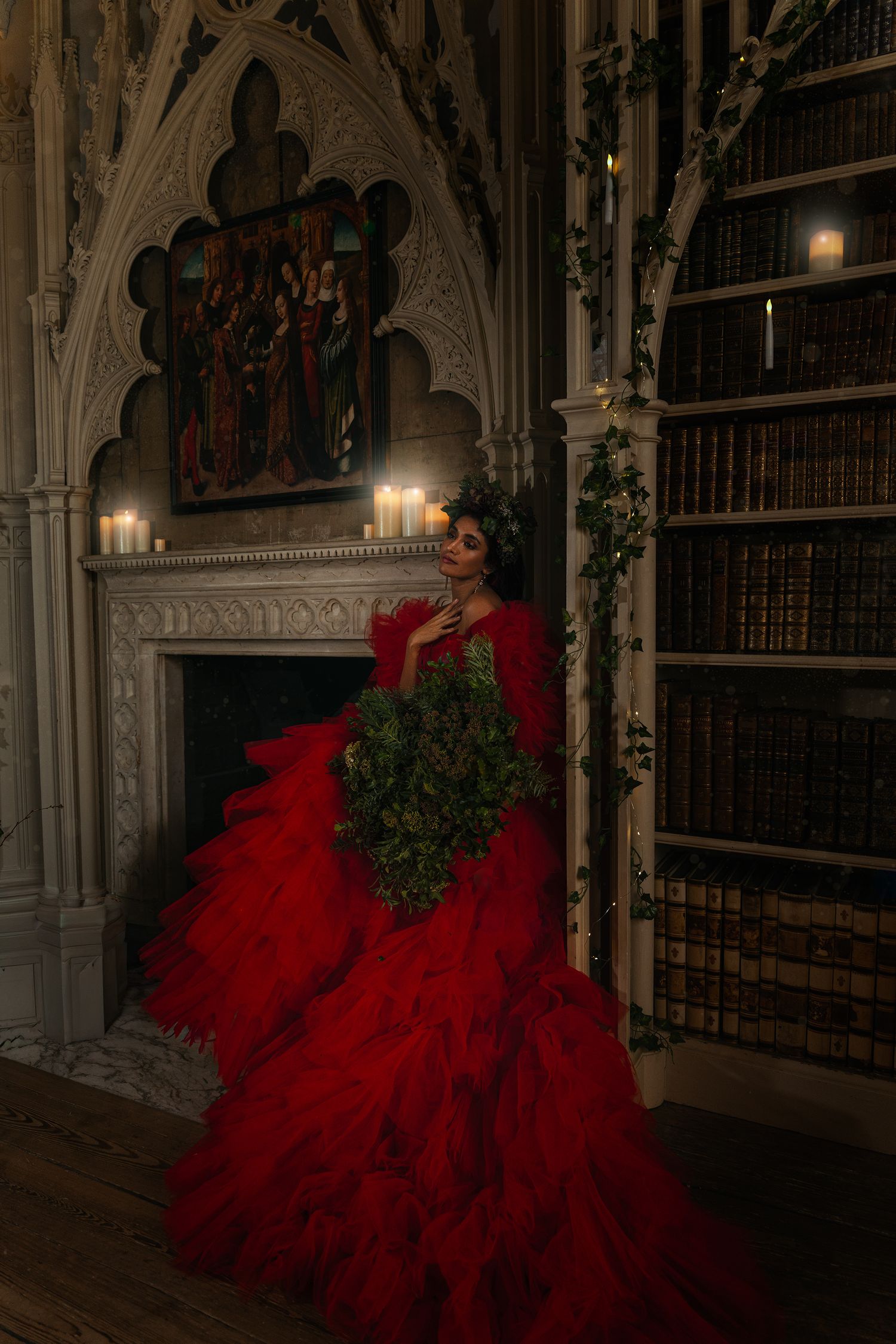 Woman in dramatic red ruffled gown holding greenery bouquet beside gothic fireplace with candles and bookshelf.