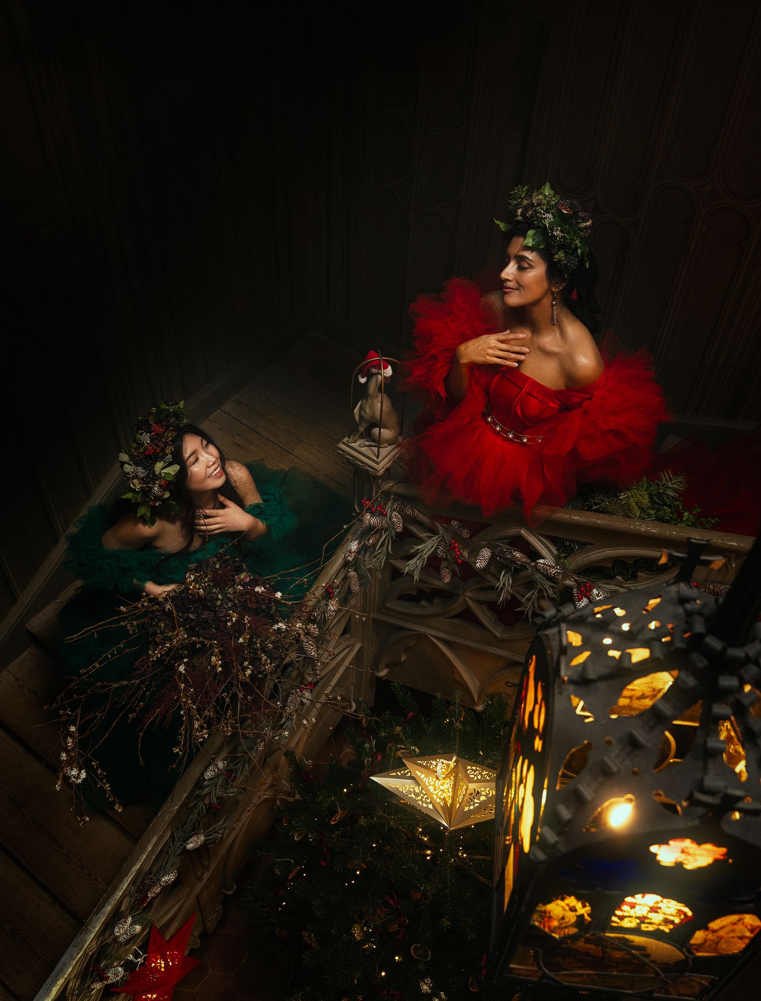 Two women in red and green gowns pose on ornate staircase with glowing star lanterns and flowers.