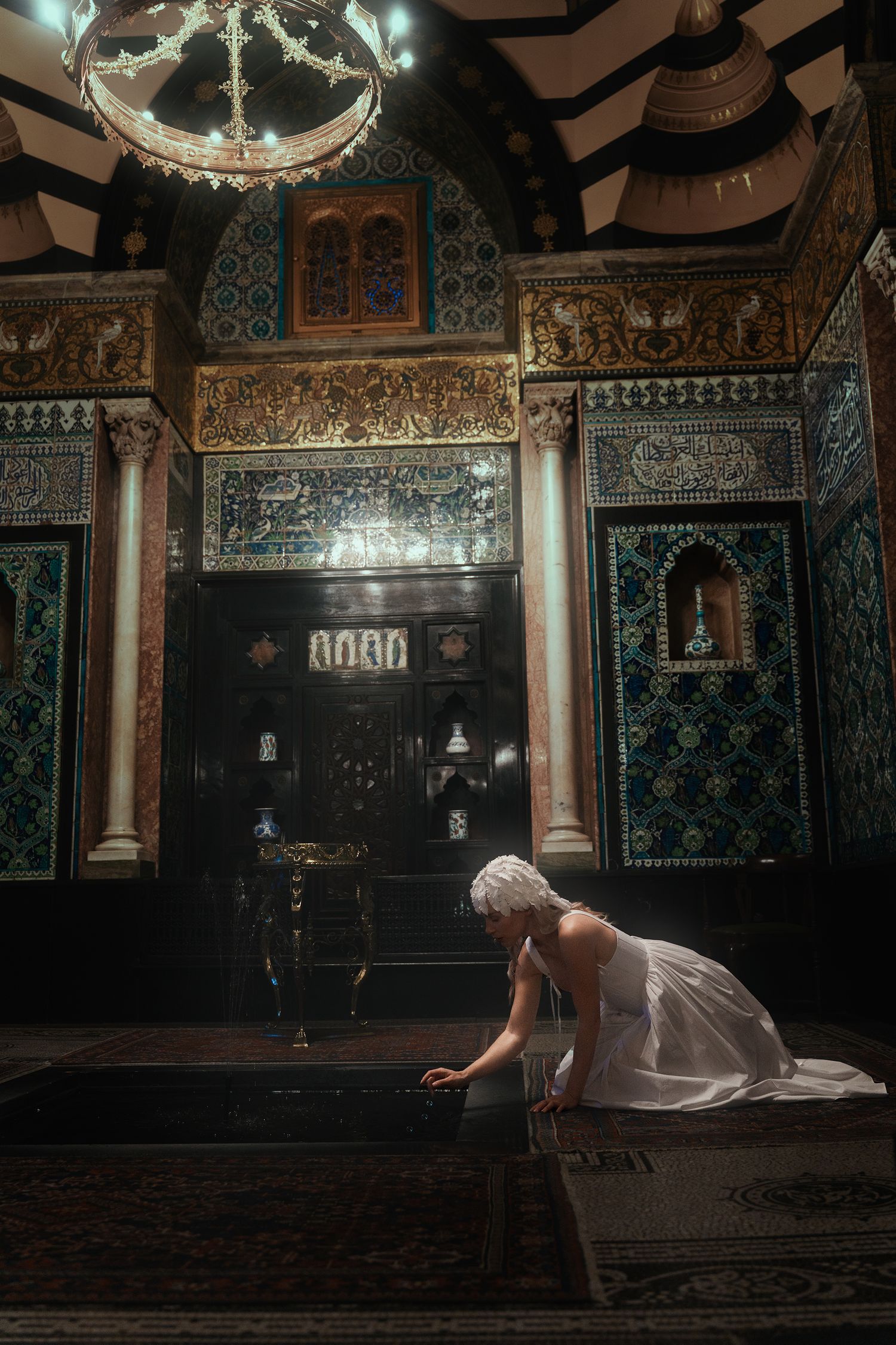 Woman in white dress kneels on ornate carpet in lavish Islamic-inspired interior with intricate tilework and chandelier above.