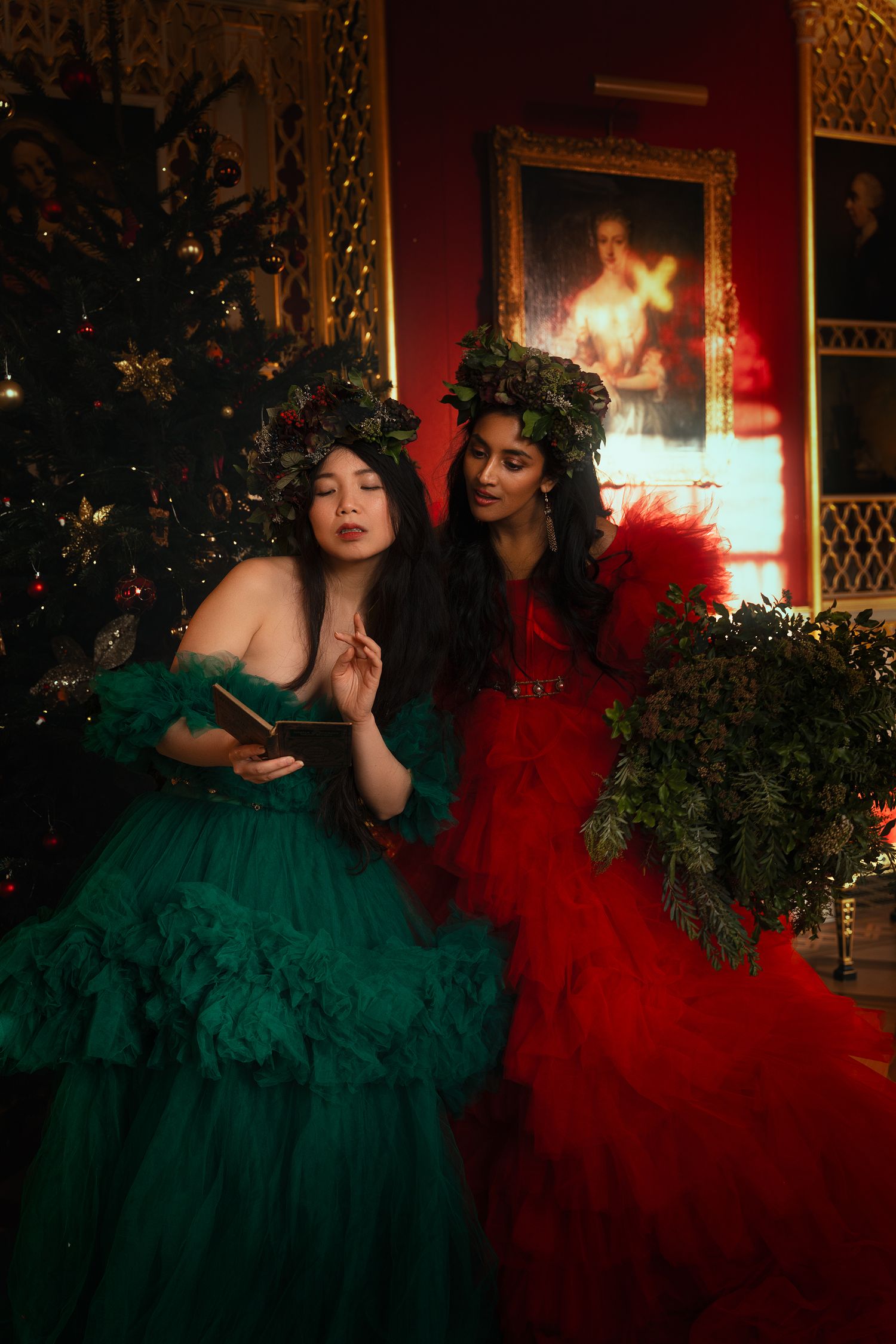 Two women in green and red gowns with floral crowns reading a book by Christmas tree.