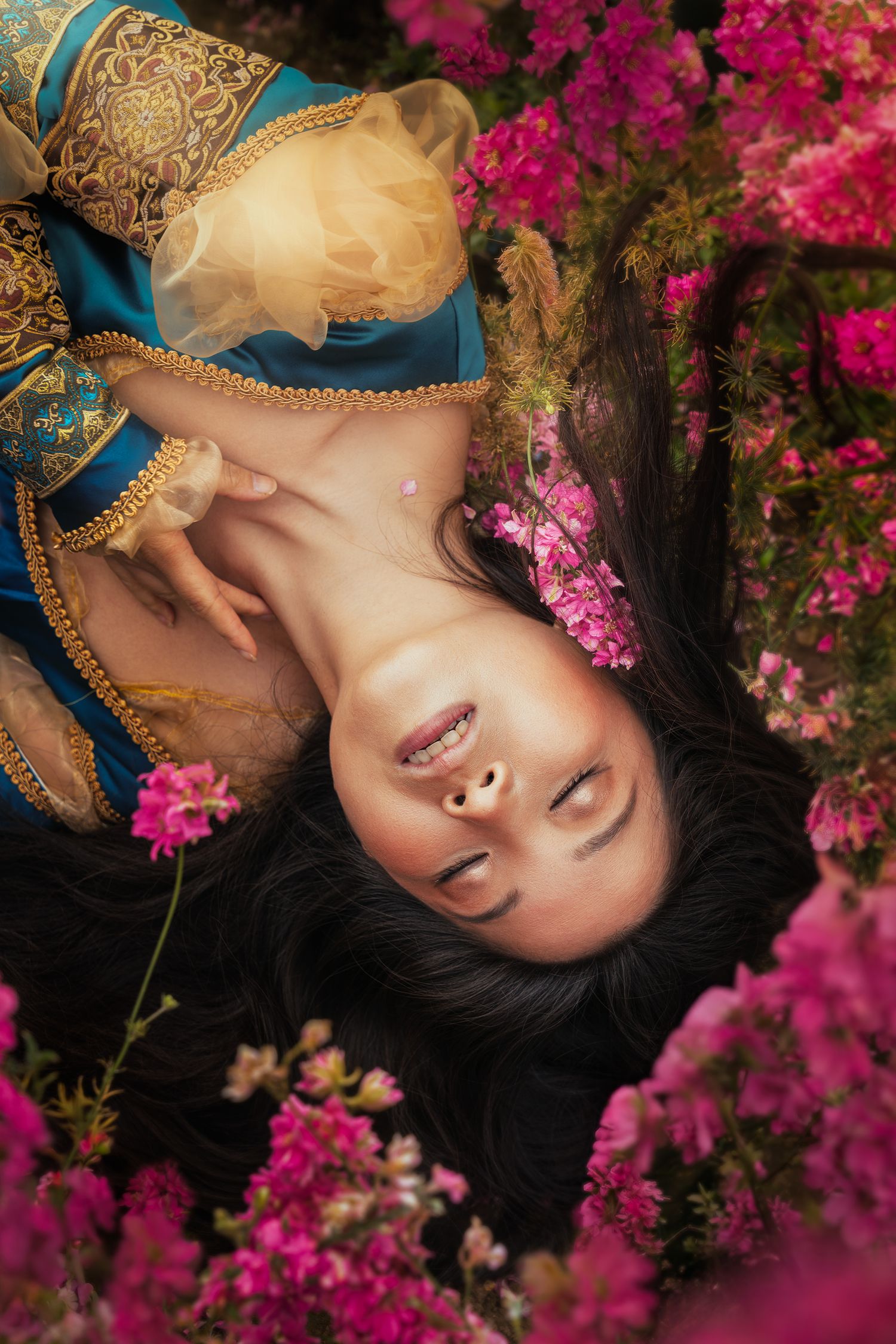 A woman in ornate blue and gold dress lies among bright pink bougainvillea flowers.
