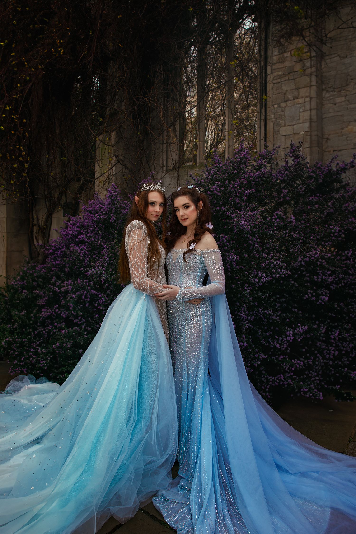Two women in elegant blue sequined and tulle fantasy gowns embrace before purple flowering stone wall backdrop.