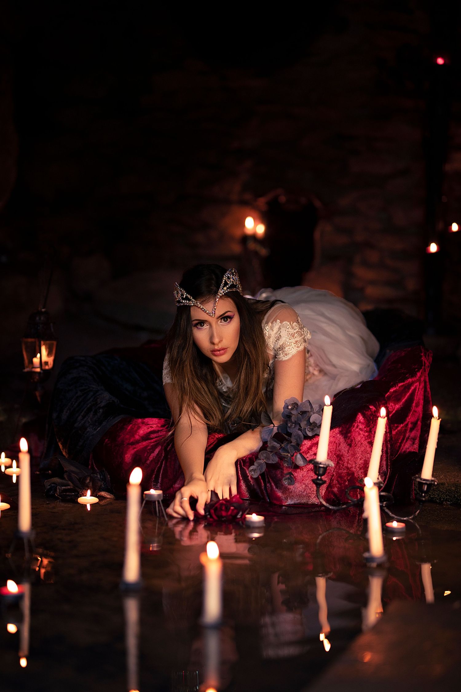 A woman in a dark fantasy queen costume with crown lies among glowing candles in a dimly lit stone chamber.
