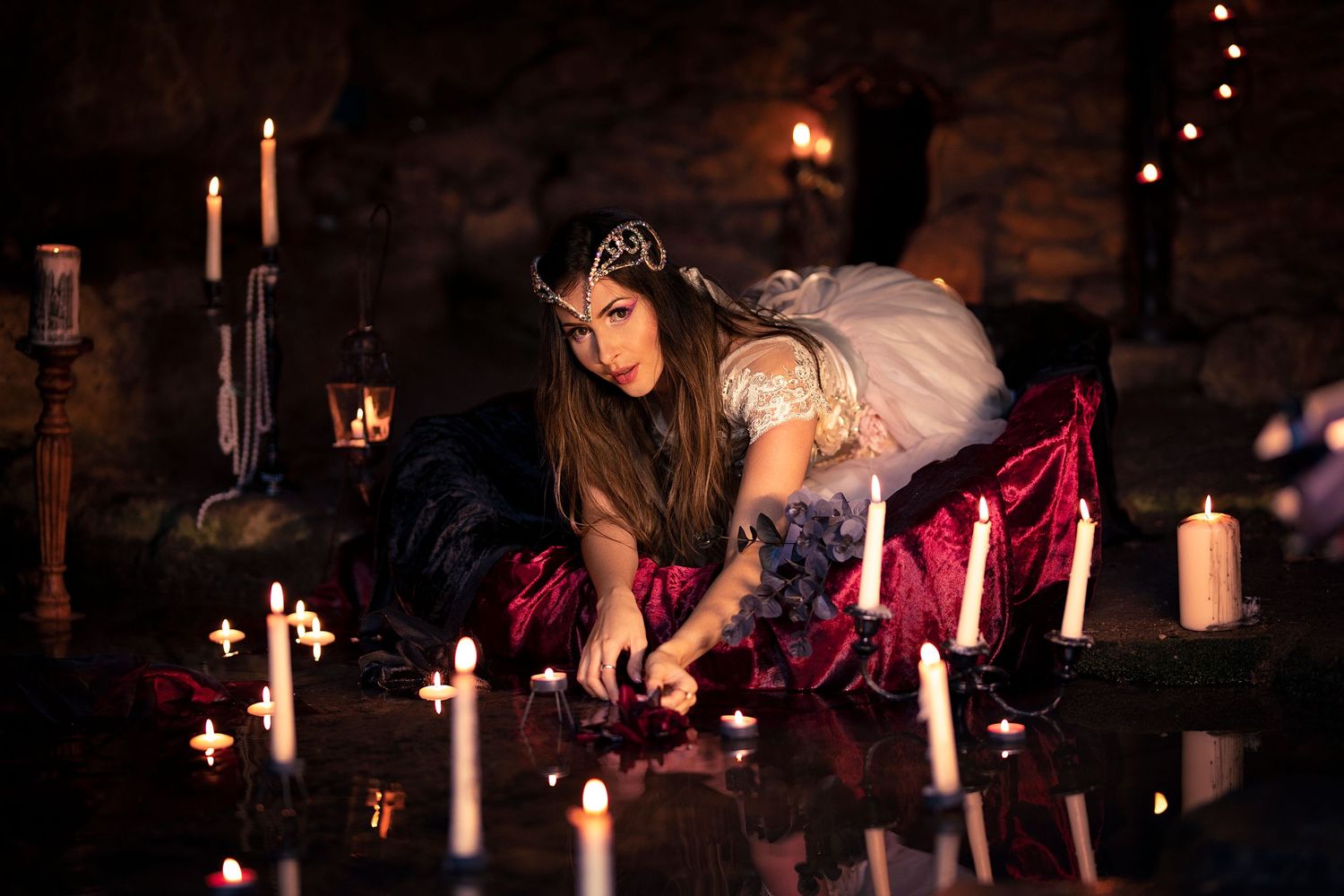 Woman in dark fantasy costume with crown reaches toward candles on reflective surface in candlelit stone room.