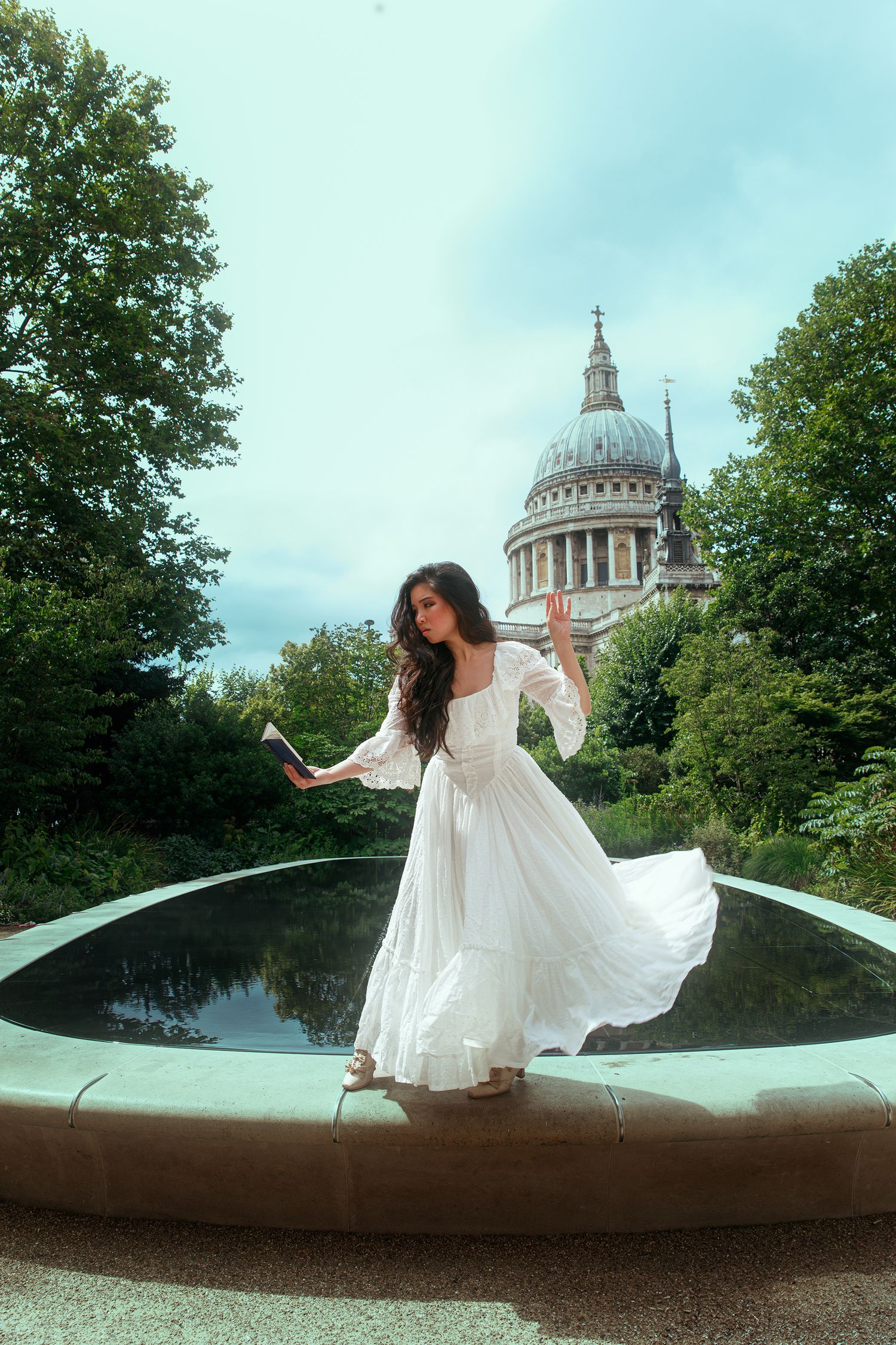 Woman in white flowing dress poses on fountain edge with St Paul's Cathedral dome visible among trees.