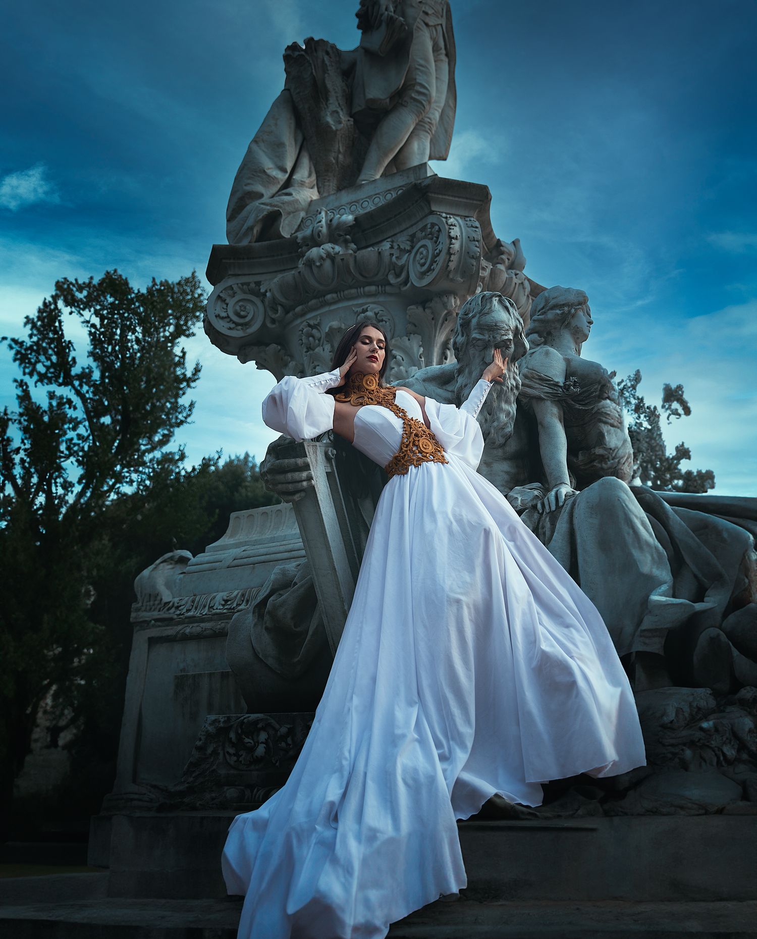 Woman in flowing white gown with gold corset posing dramatically at classical stone monument.