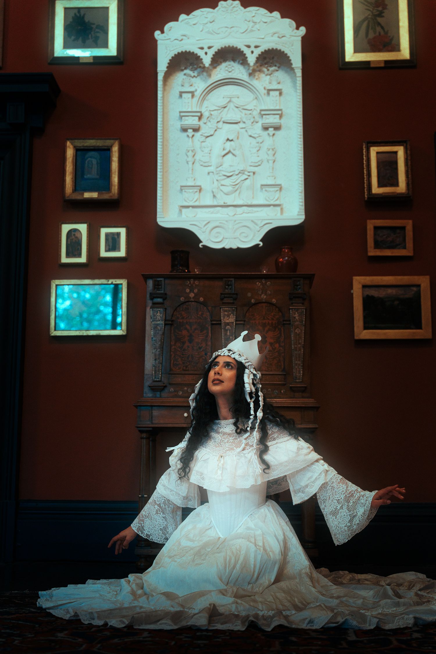 Woman in white lace Victorian dress and crown seated before ornate carved wall panel.