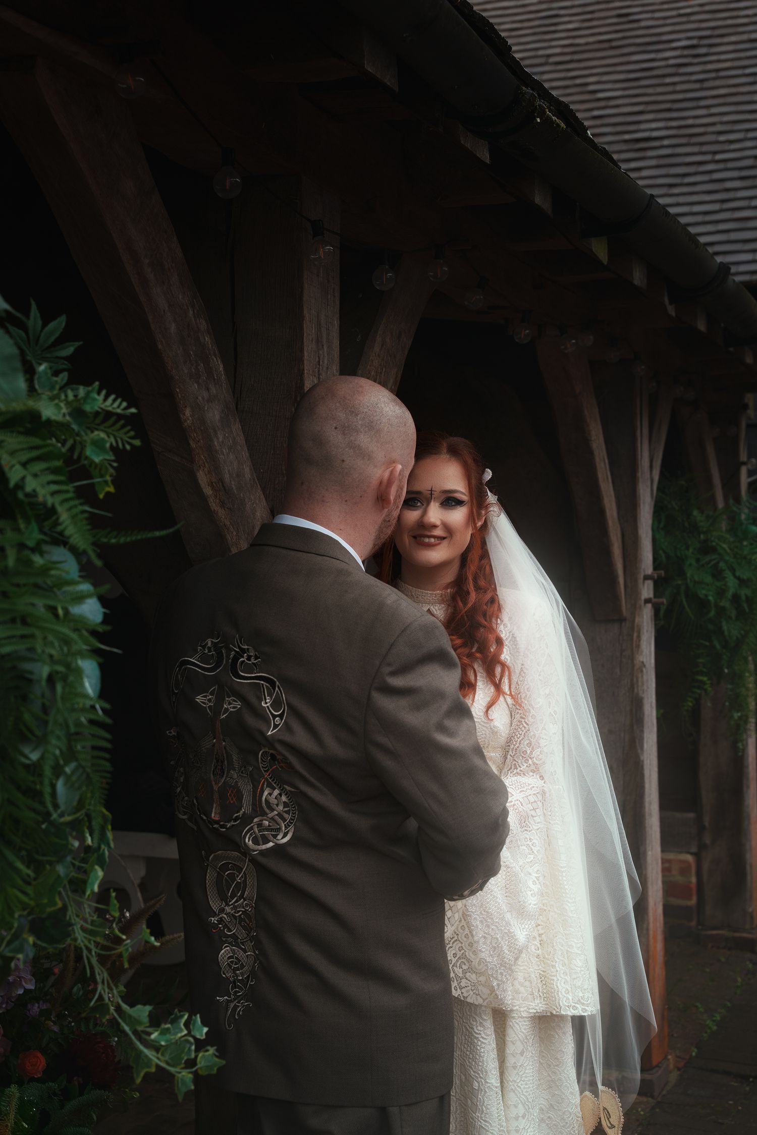 A smiling red-haired bride in a lace wedding gown and veil stands with her groom at a rustic venue.