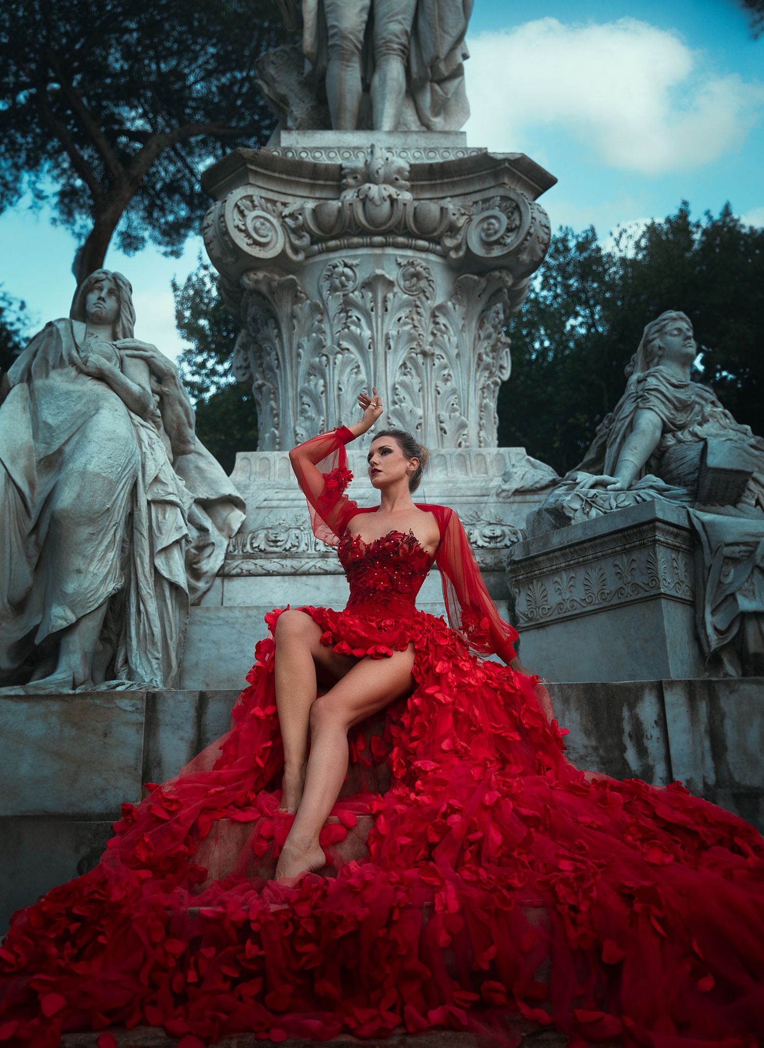 Woman in dramatic red floral gown posing on ornate stone monument with sculptures.
