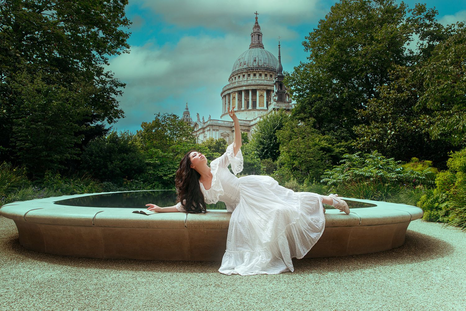 Woman in white dress reclines gracefully in stone fountain with St Pauls Cathedral in background.