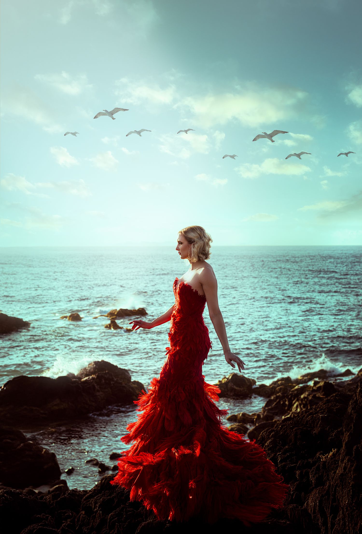 A woman in a dramatic red ruffled gown stands on rocky coastal cliffs with birds soaring overhead.