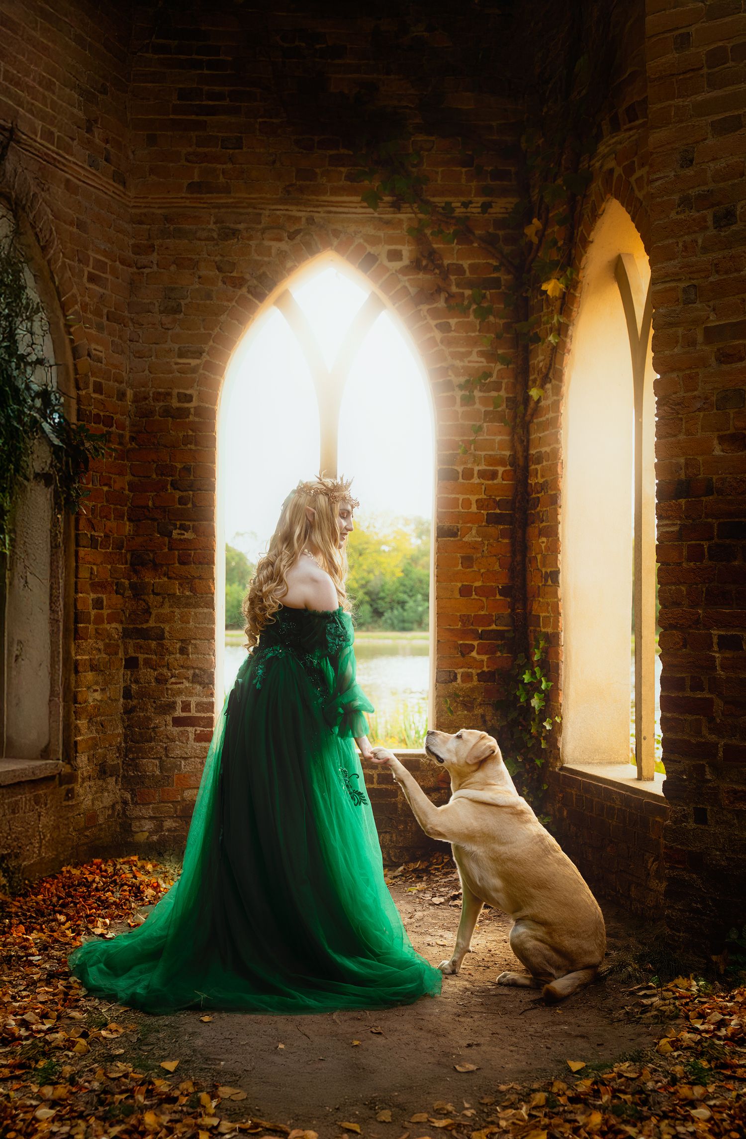 Blonde woman in emerald green tulle gown interacts with golden Labrador in sunlit brick archway ruins.