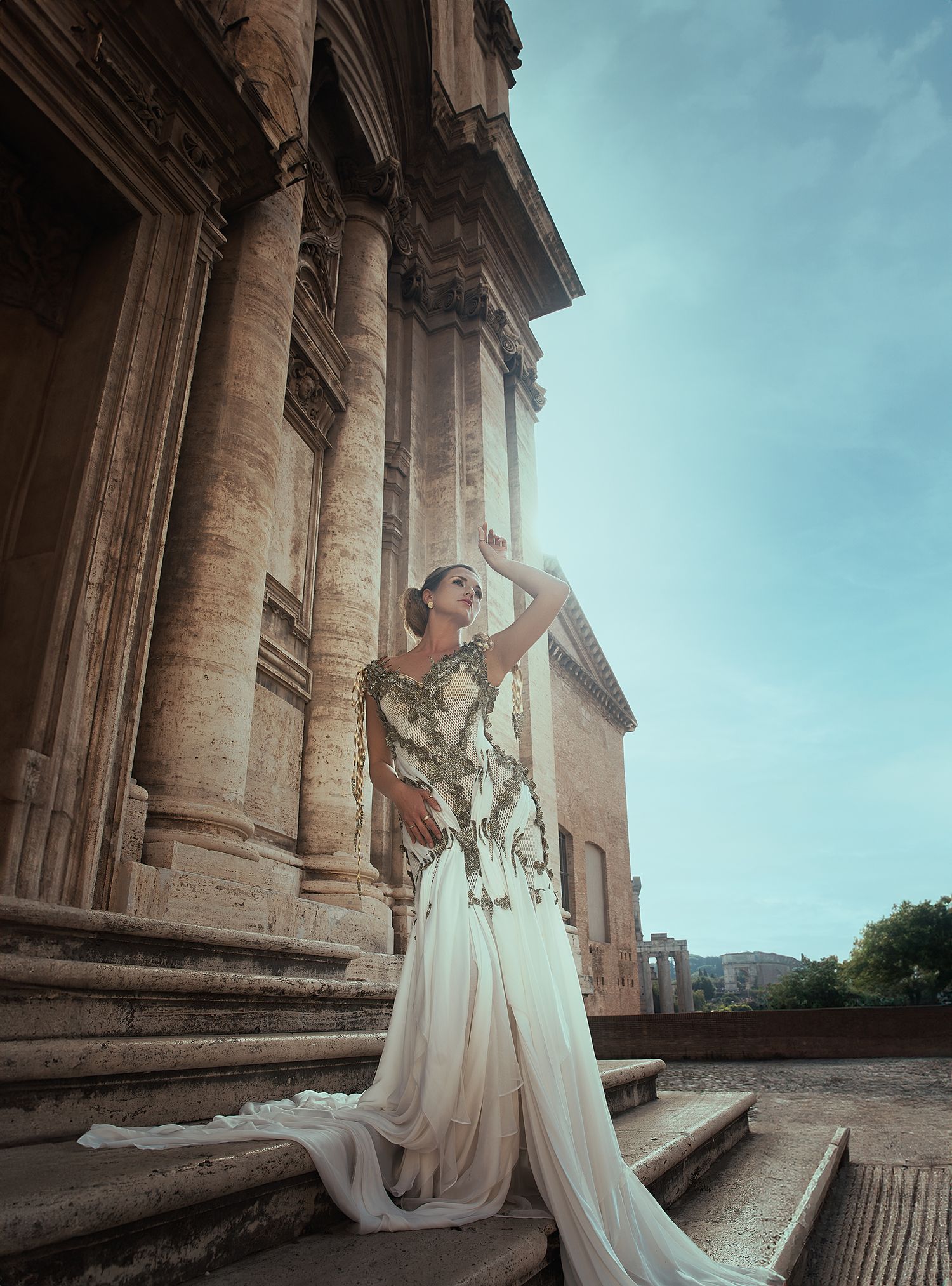 Elegant woman in embroidered white gown posing dramatically on ancient Roman temple steps.