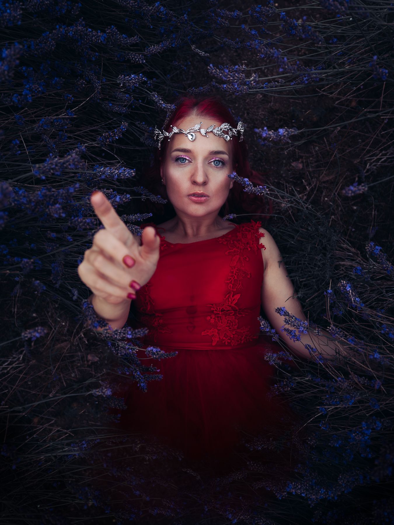 Woman in red dress and silver crown pointing finger, surrounded by purple lavender flowers in dark setting.