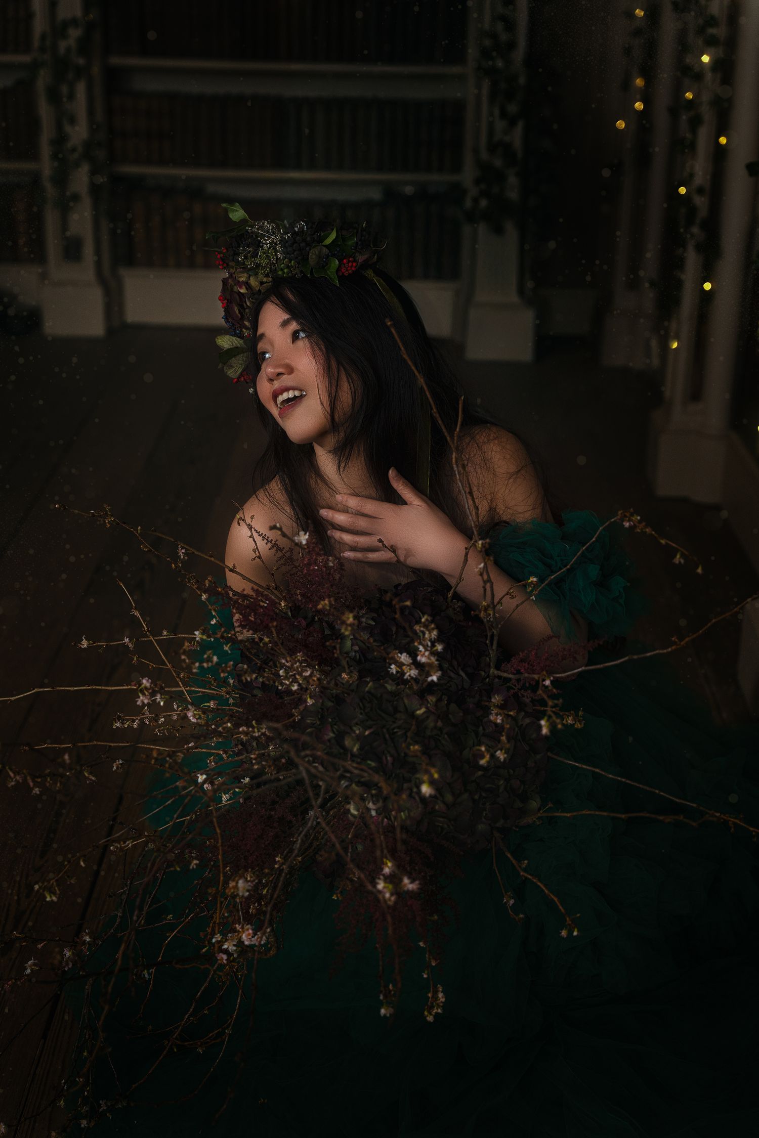Smiling woman in emerald green gown with floral crown holds branches in a dimly lit ornate interior.