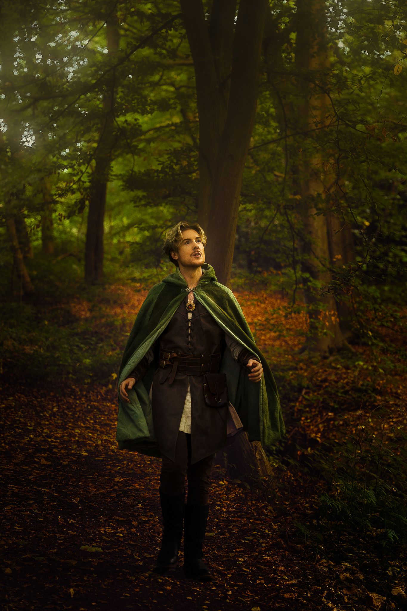 Young man in green fantasy cloak standing on forest path in atmospheric woodland setting.