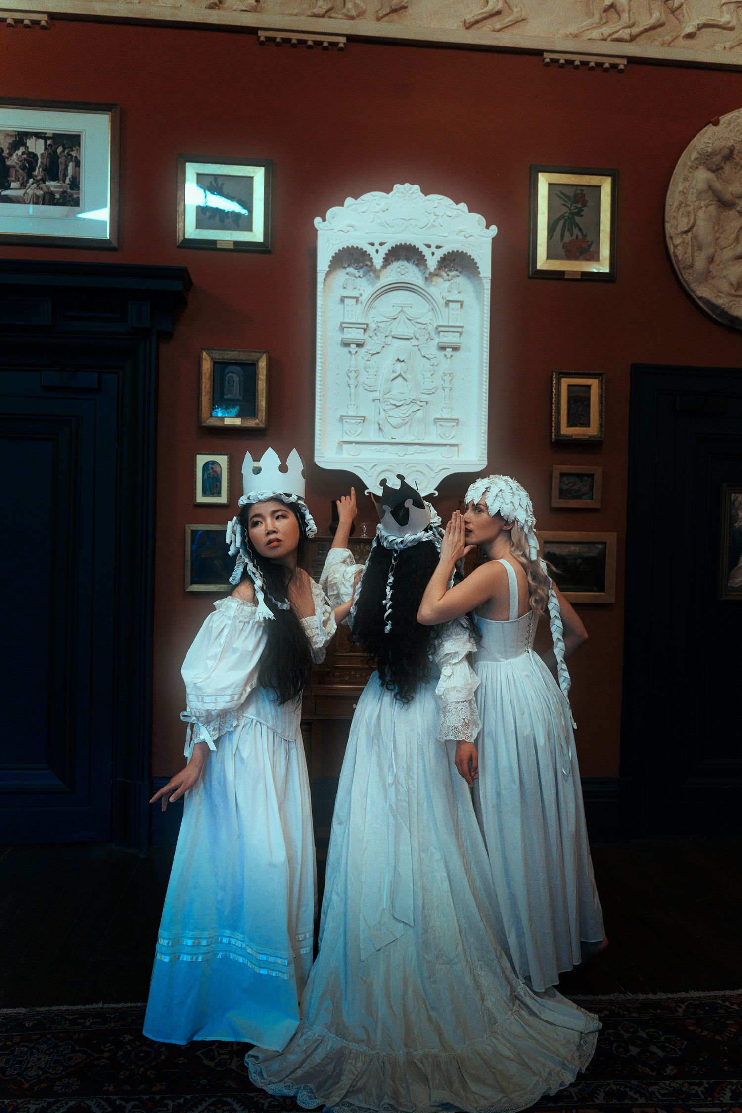 Three women in white Victorian dresses and crowns standing before ornate carved wall panel.