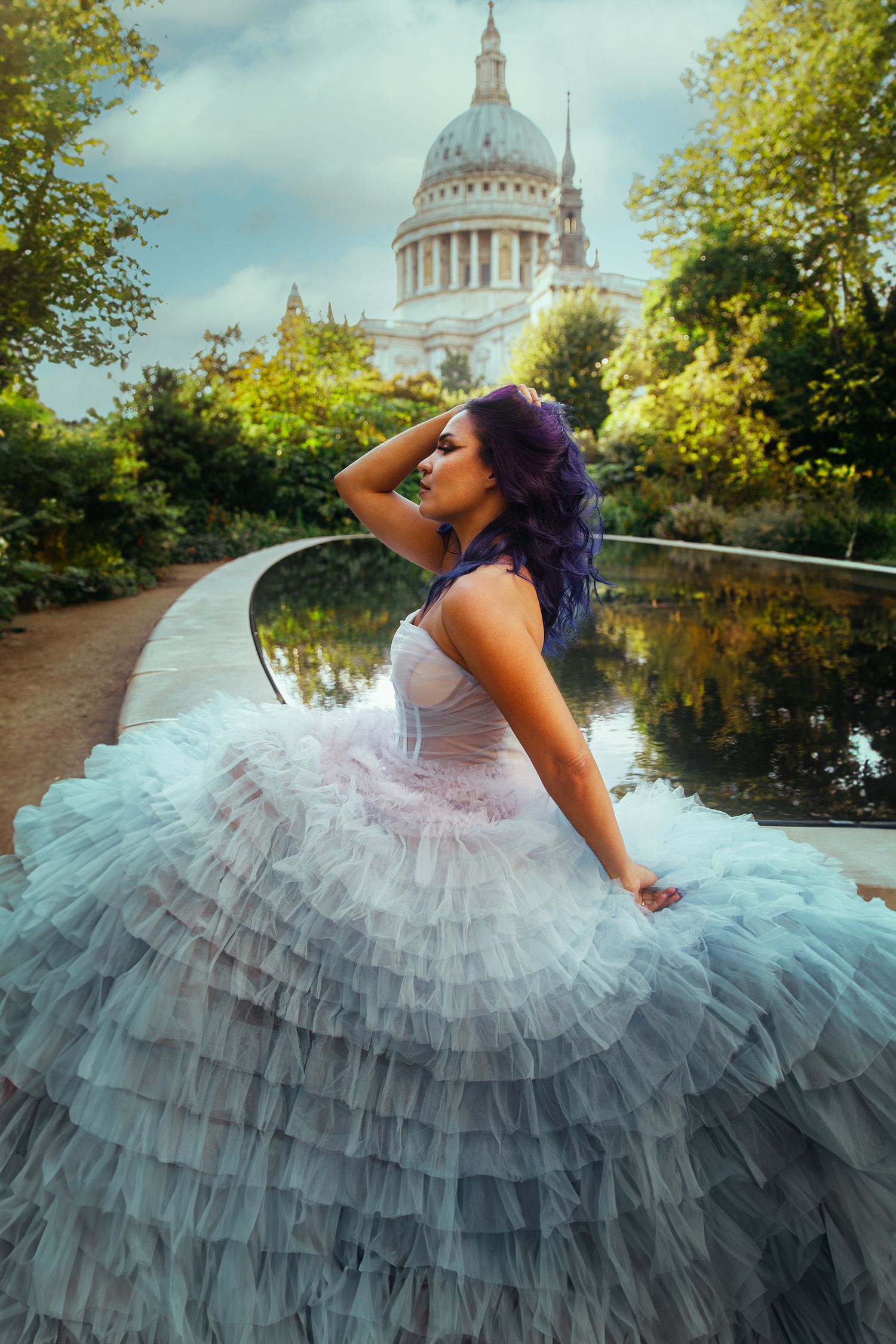 Woman with purple hair in ruffled white ballgown posing near pond with domed cathedral.