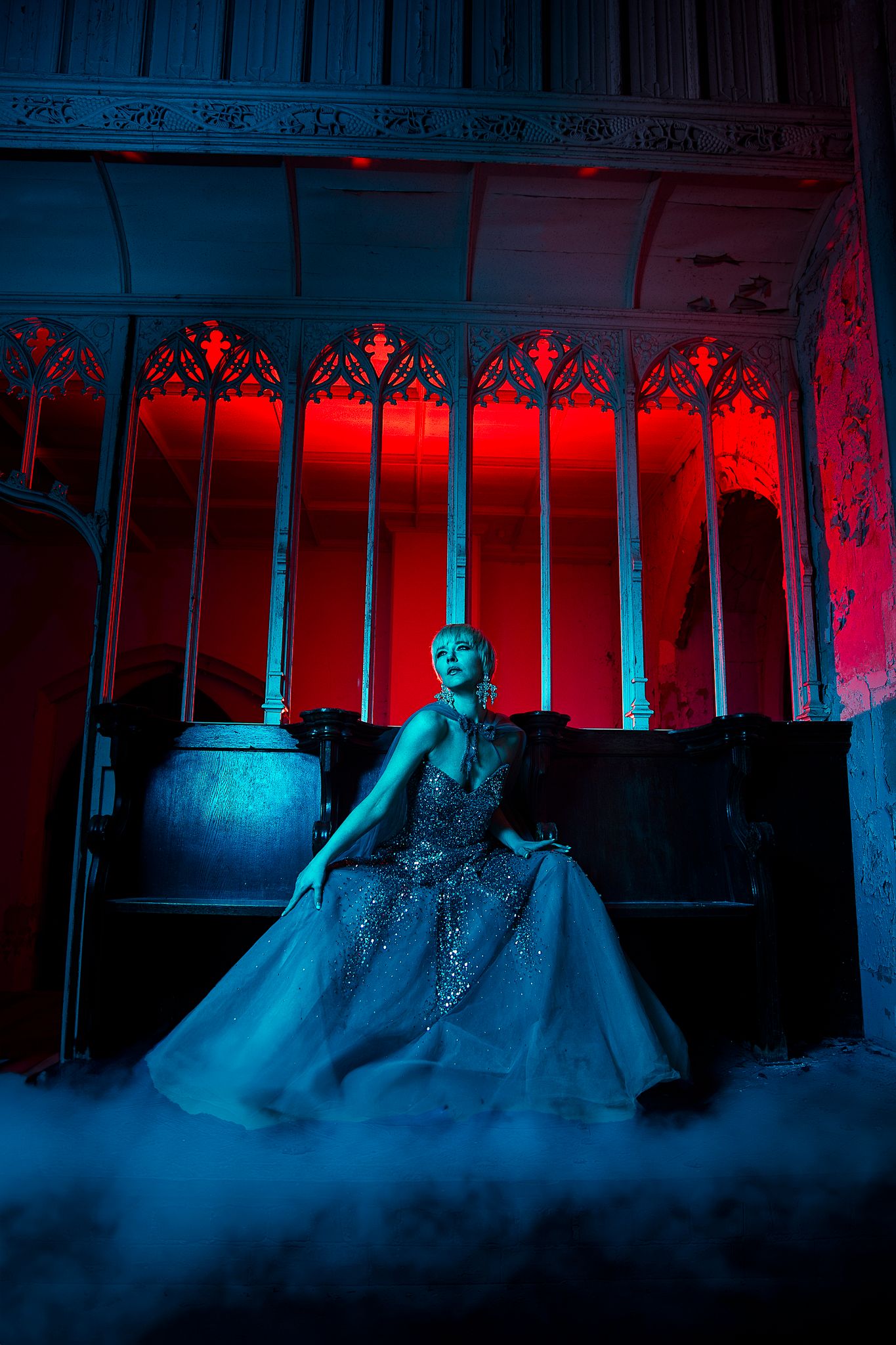 Woman in sparkling silver ball gown sits in gothic interior with red and blue dramatic lighting and mist.