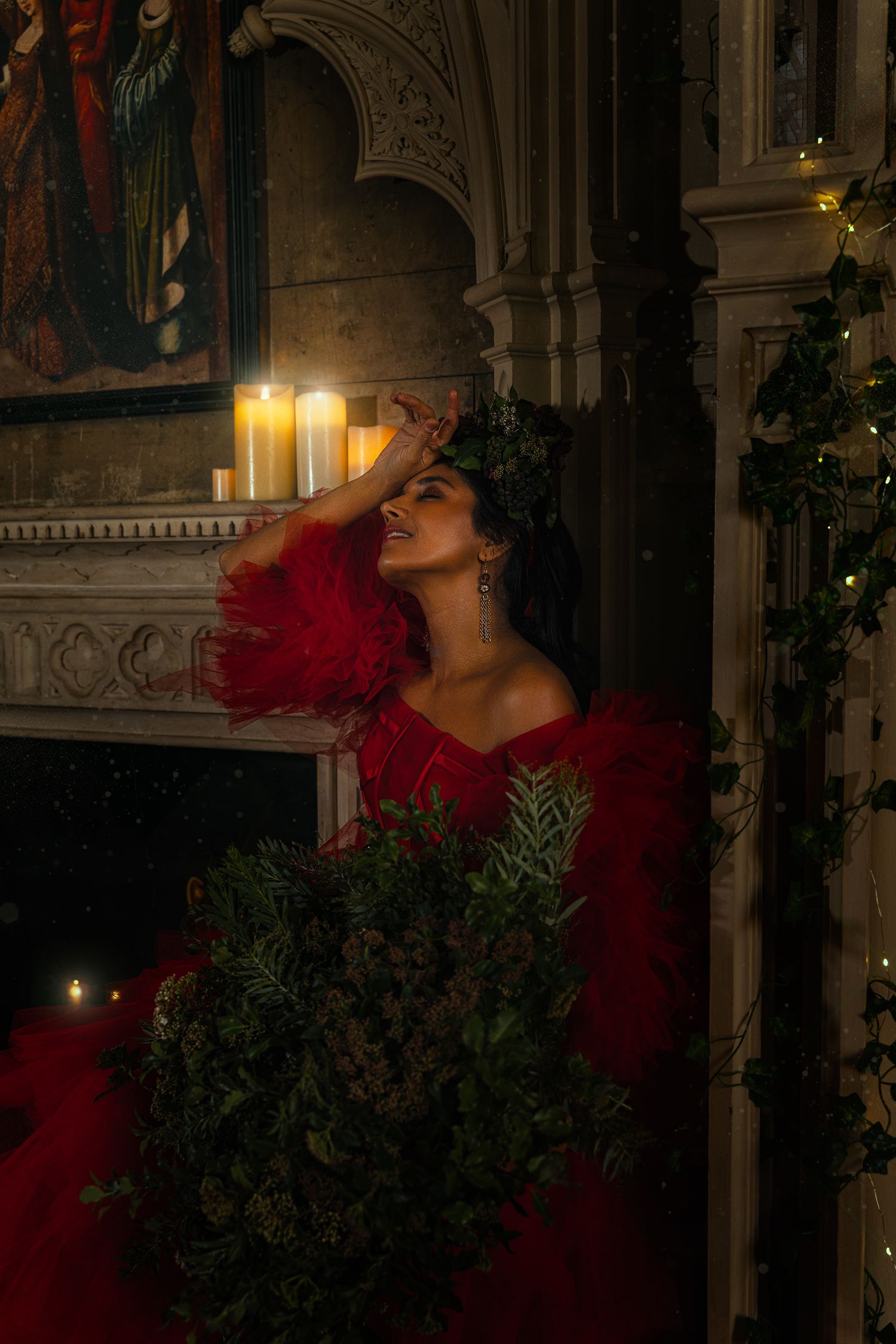 Woman in red feathered gown holding greenery beside candlelit Gothic fireplace with ivy decor.