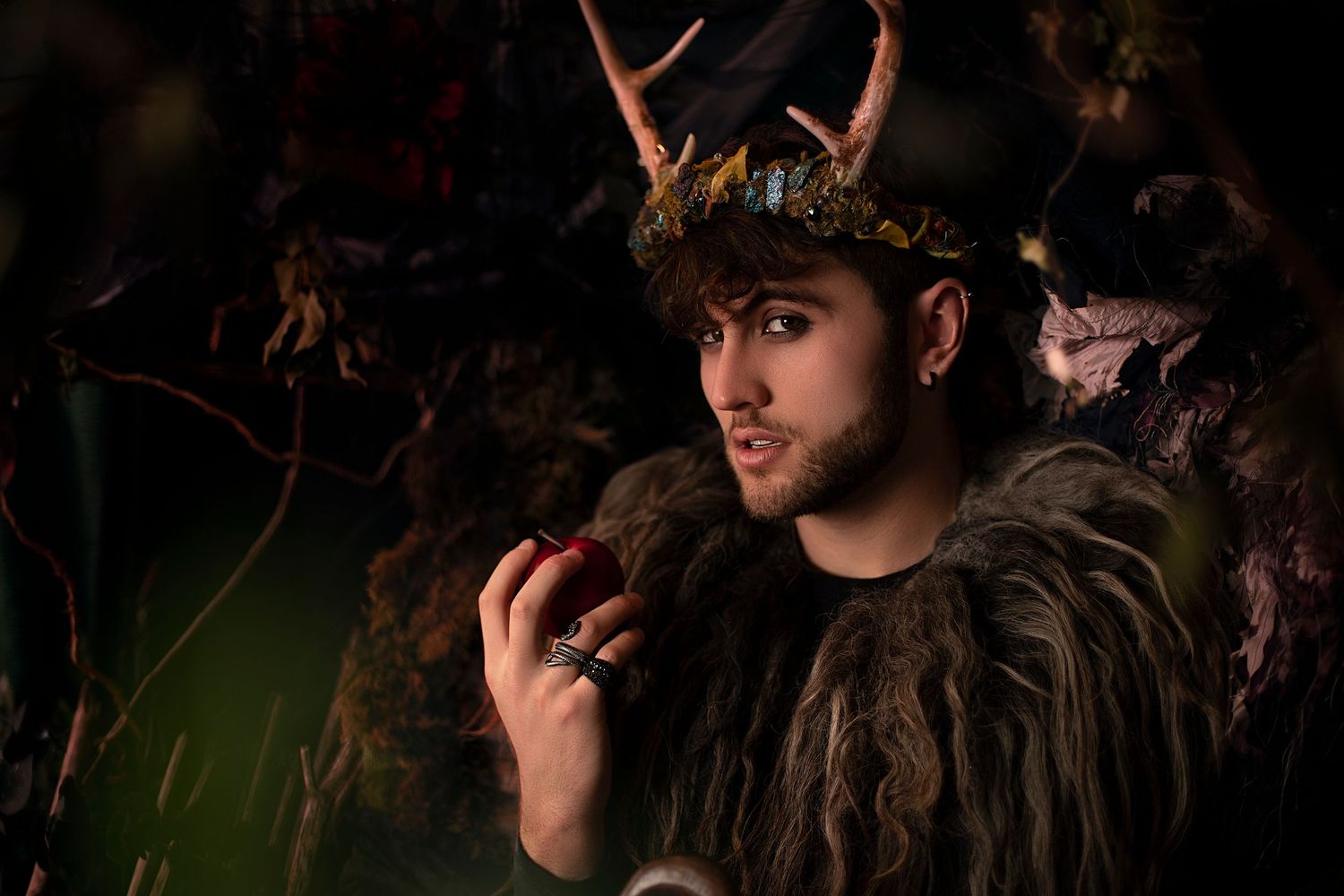 Dark-haired man wearing antler crown and fur costume holds red apple in moody forest fantasy setting.