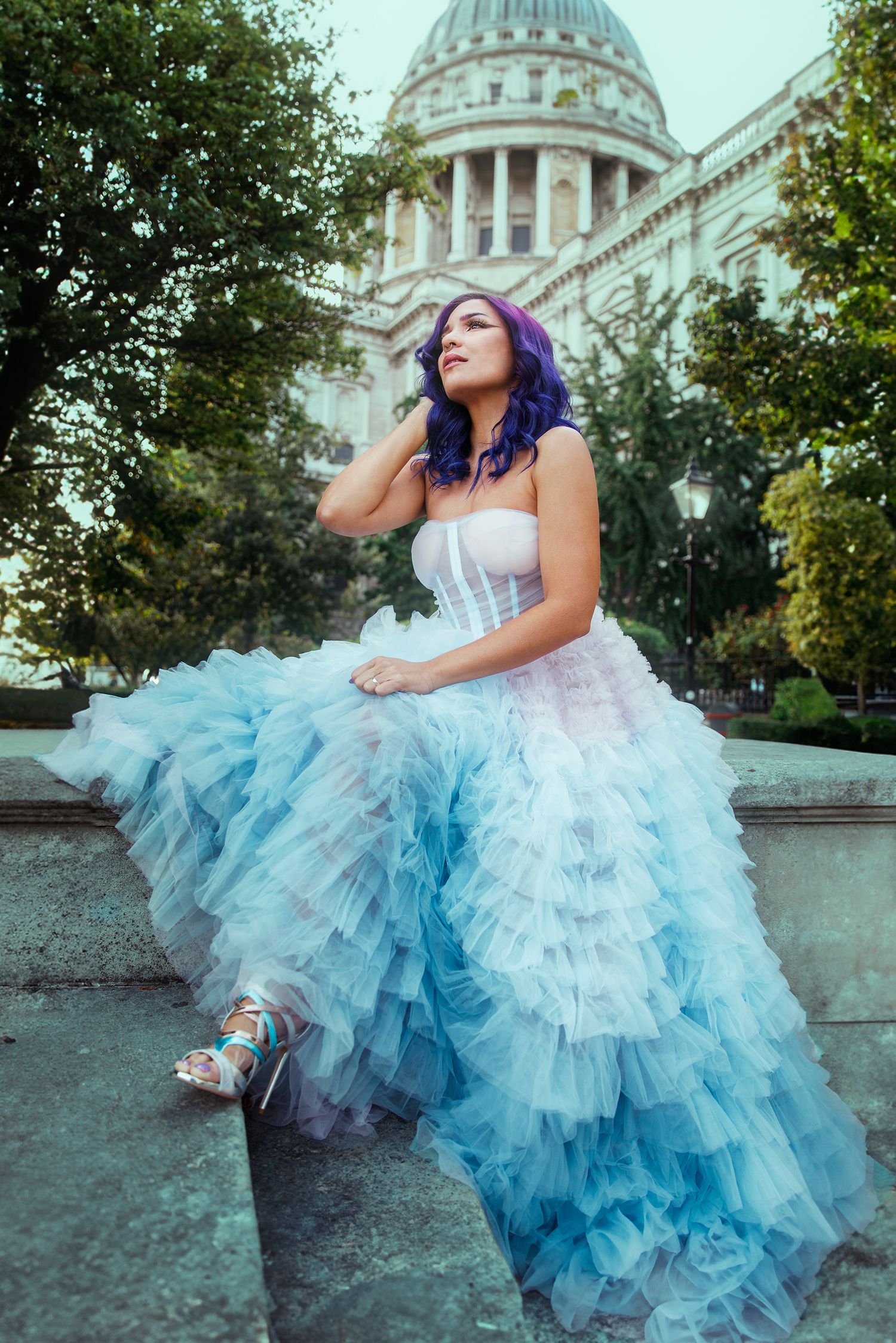 A woman with purple hair in a blue ruffled tulle gown sits on stone steps before a grand domed building.