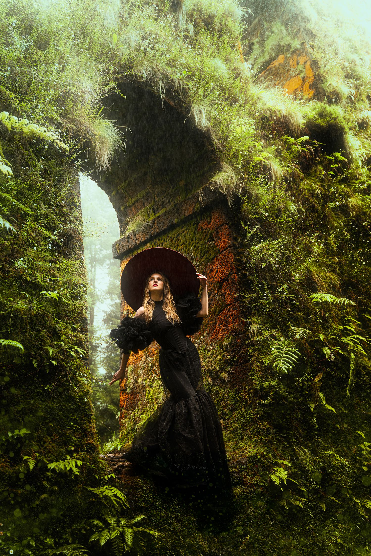 Woman in black Victorian dress and hat stands near mossy ruins with a misty waterfall.