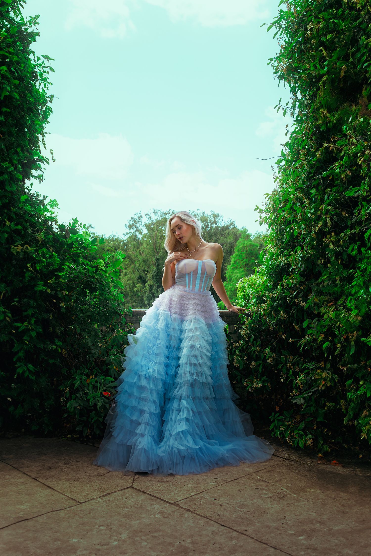 Blonde woman in blue ruffled tulle ball gown stands between tall green hedges in garden.