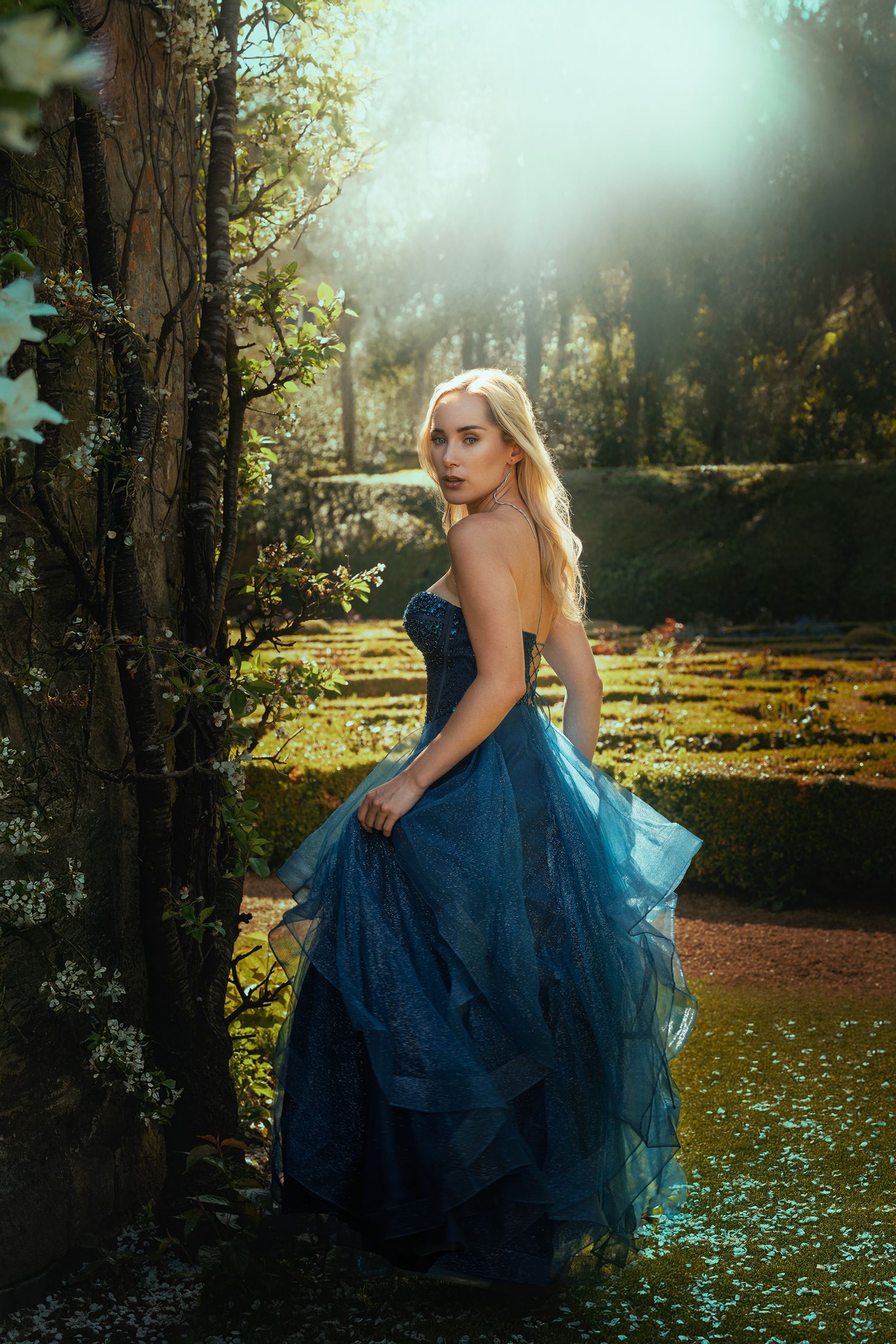 A blonde woman in a layered blue tulle ball gown stands by a tree in a dreamy sunlit garden.