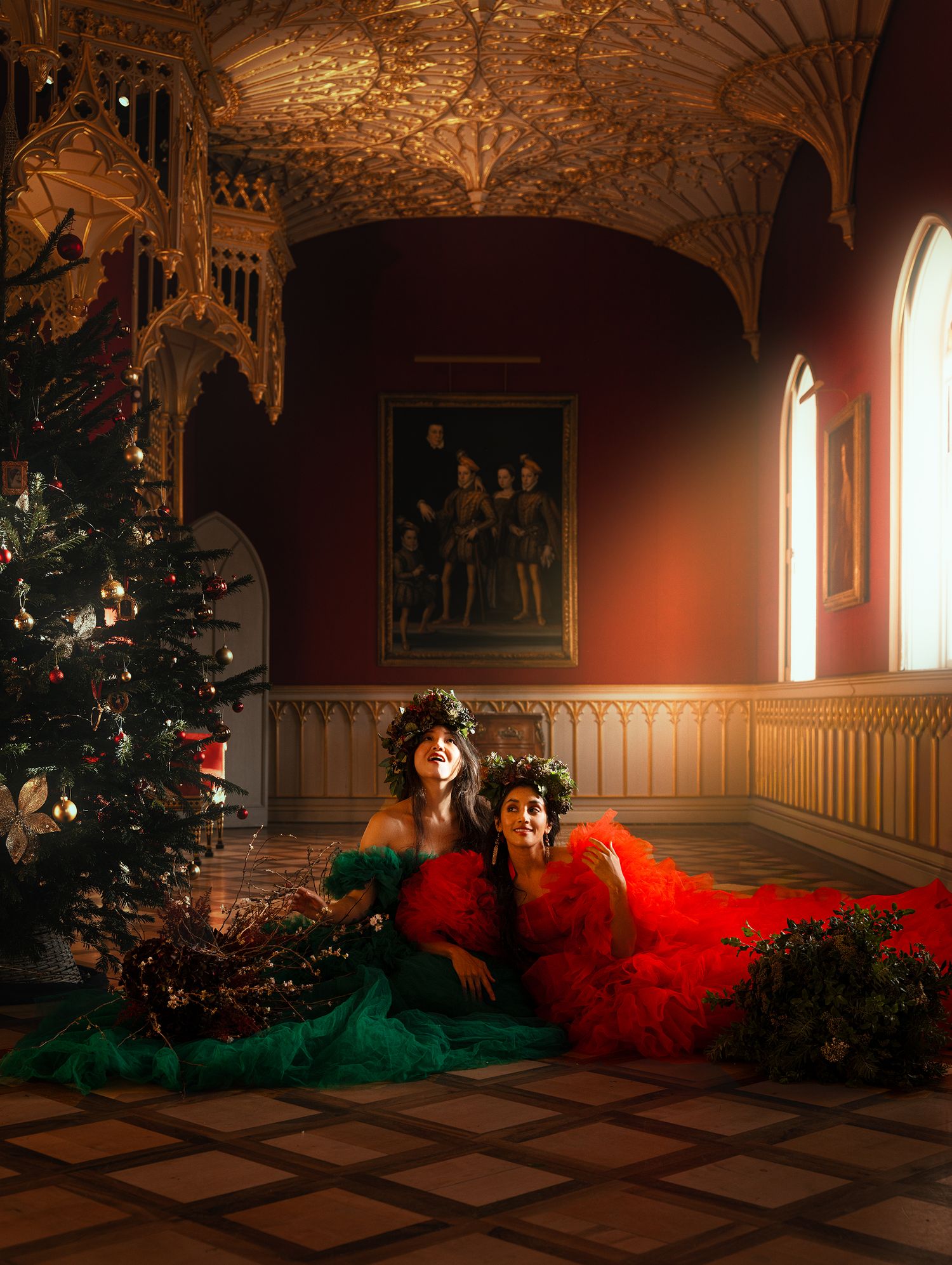 Two women in red and green gowns resting on floor beside Christmas tree in grand Gothic hall.