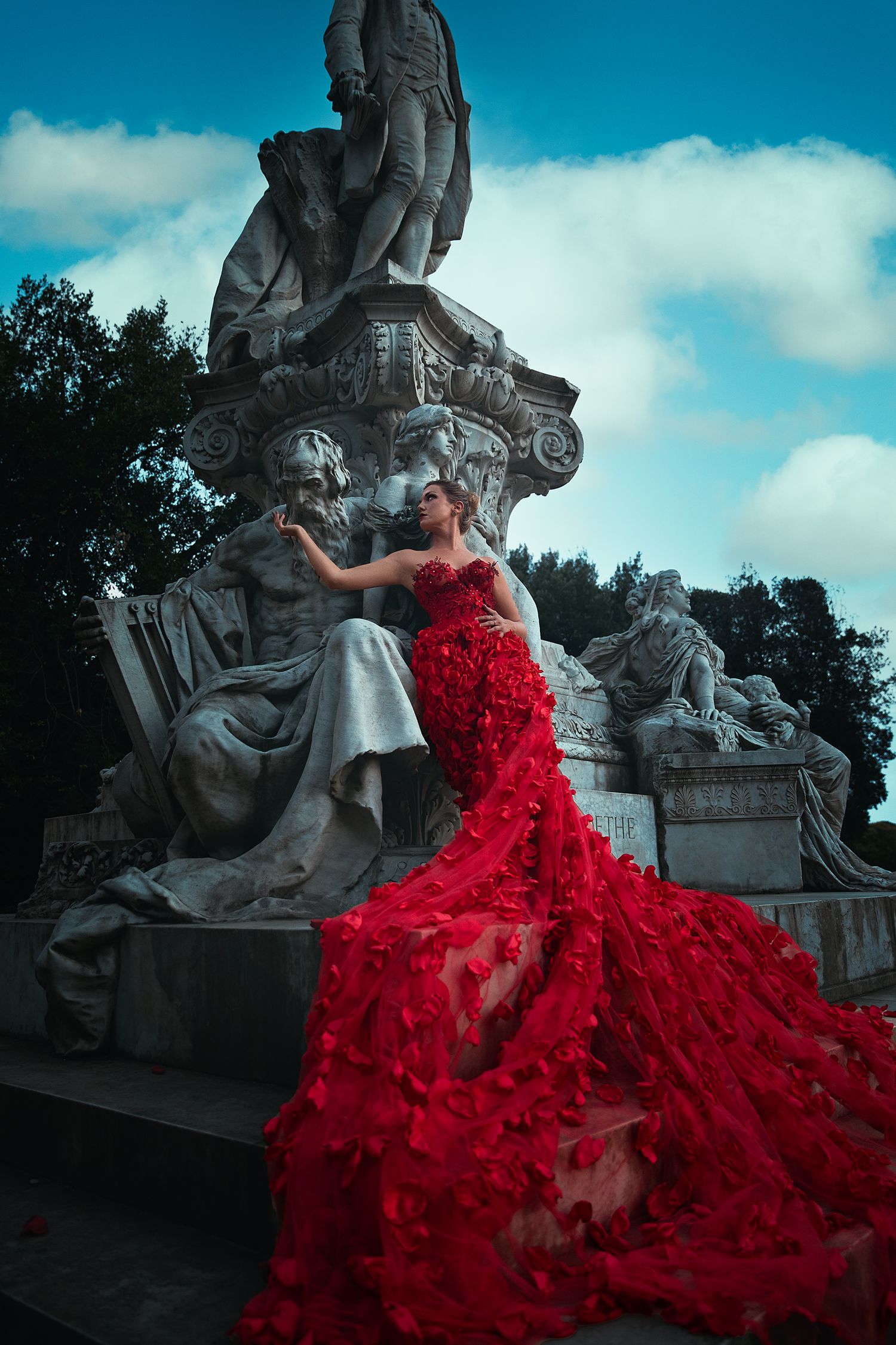 Woman in stunning red floral mermaid gown posing beside classical stone sculptures outdoors.