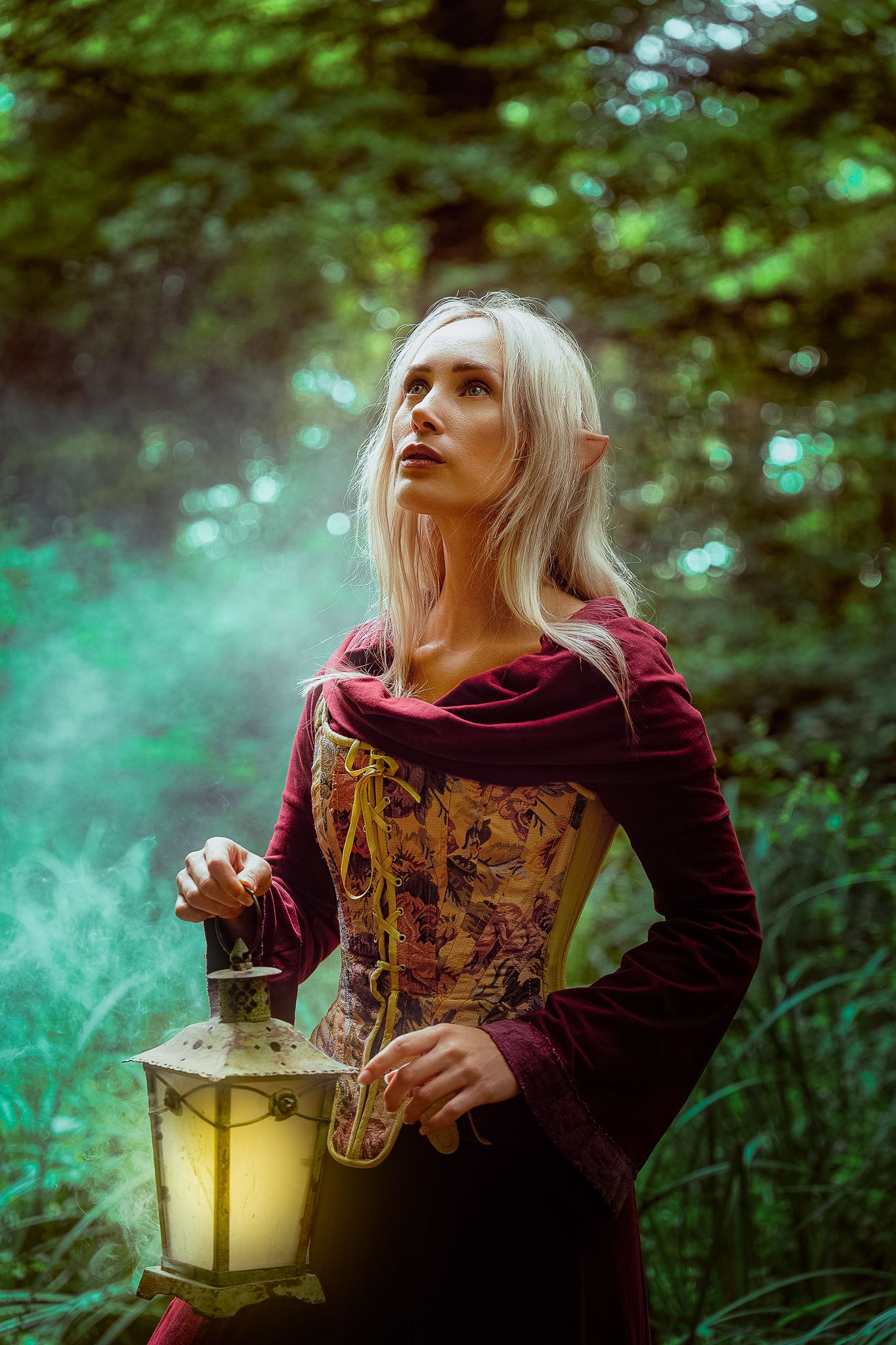 Blonde elf cosplay woman holding glowing lantern in misty enchanted forest with green light.
