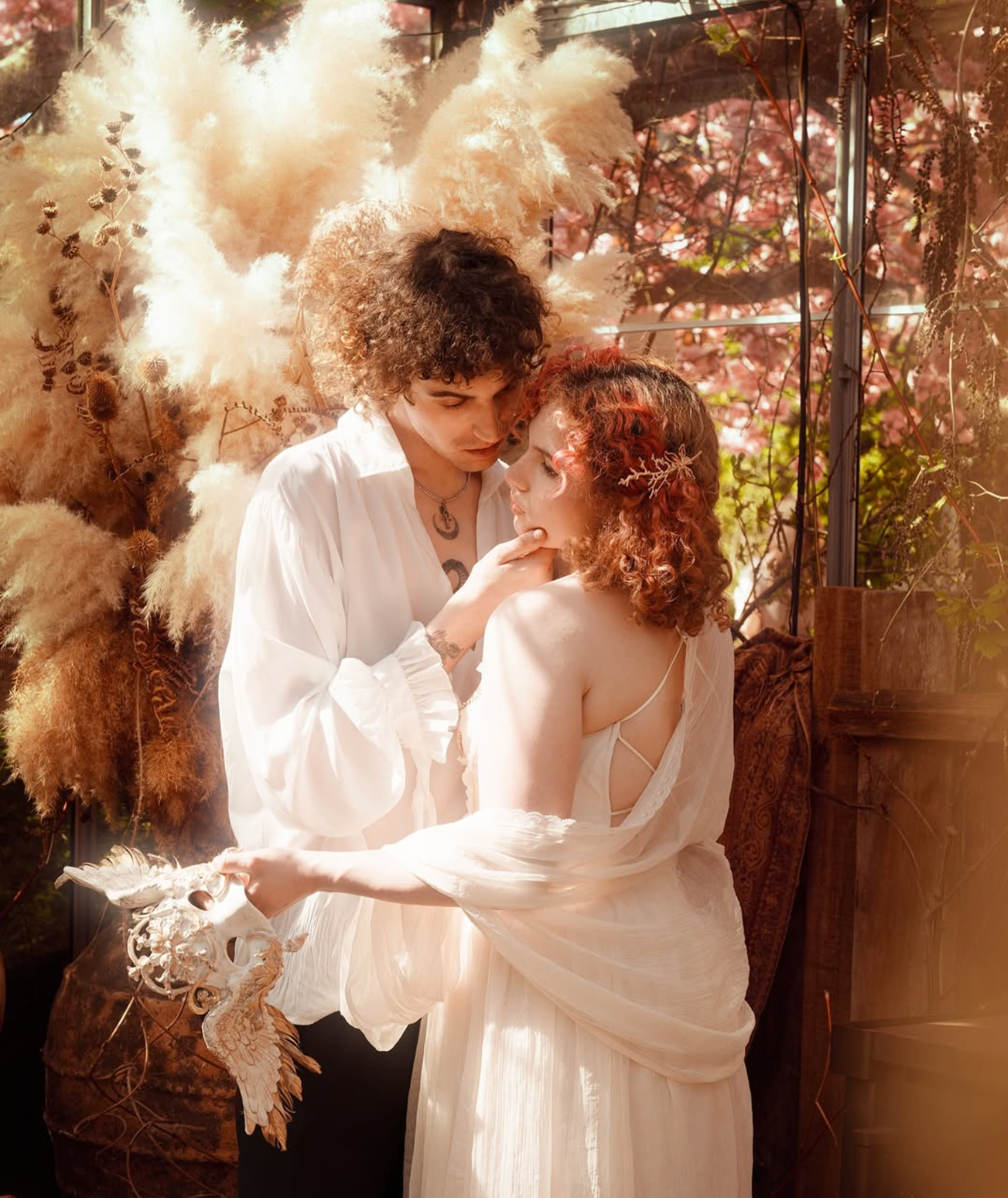 A romantic couple in white bohemian attire embraces among dried pampas grass in a dreamy outdoor setting.