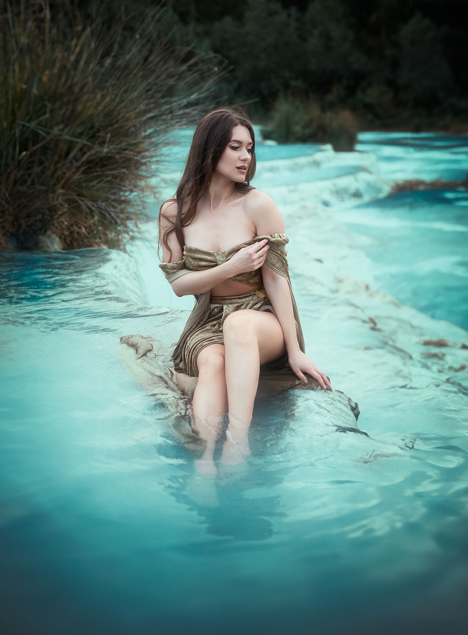 Brunette woman in gold outfit sitting on rocks with feet in turquoise thermal waters.