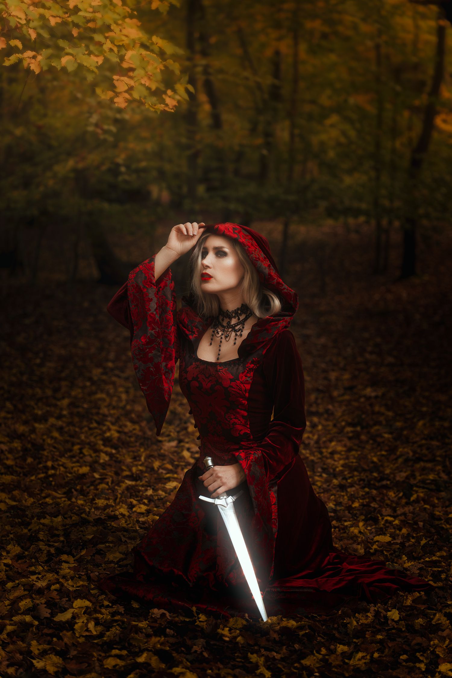 A woman in red velvet hooded gown kneels in autumn forest holding a glowing magical dagger.