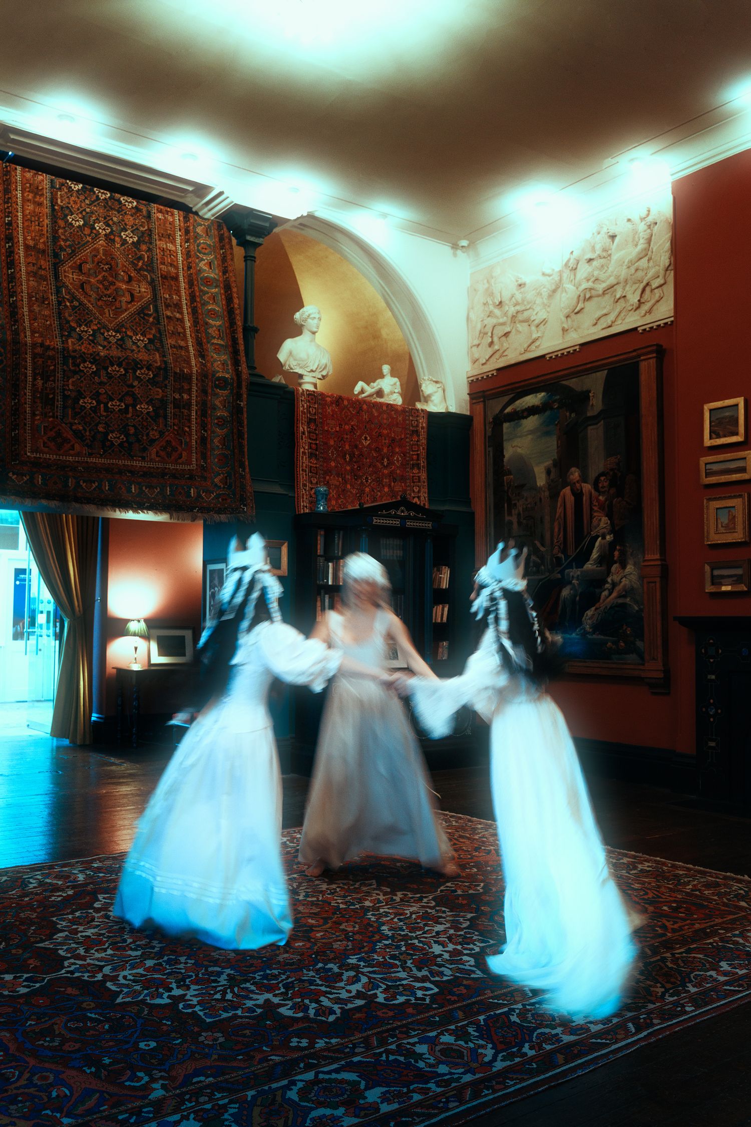 Three figures in white gowns create a motion-blur effect while dancing in an ornate room with tapestries and classical art.