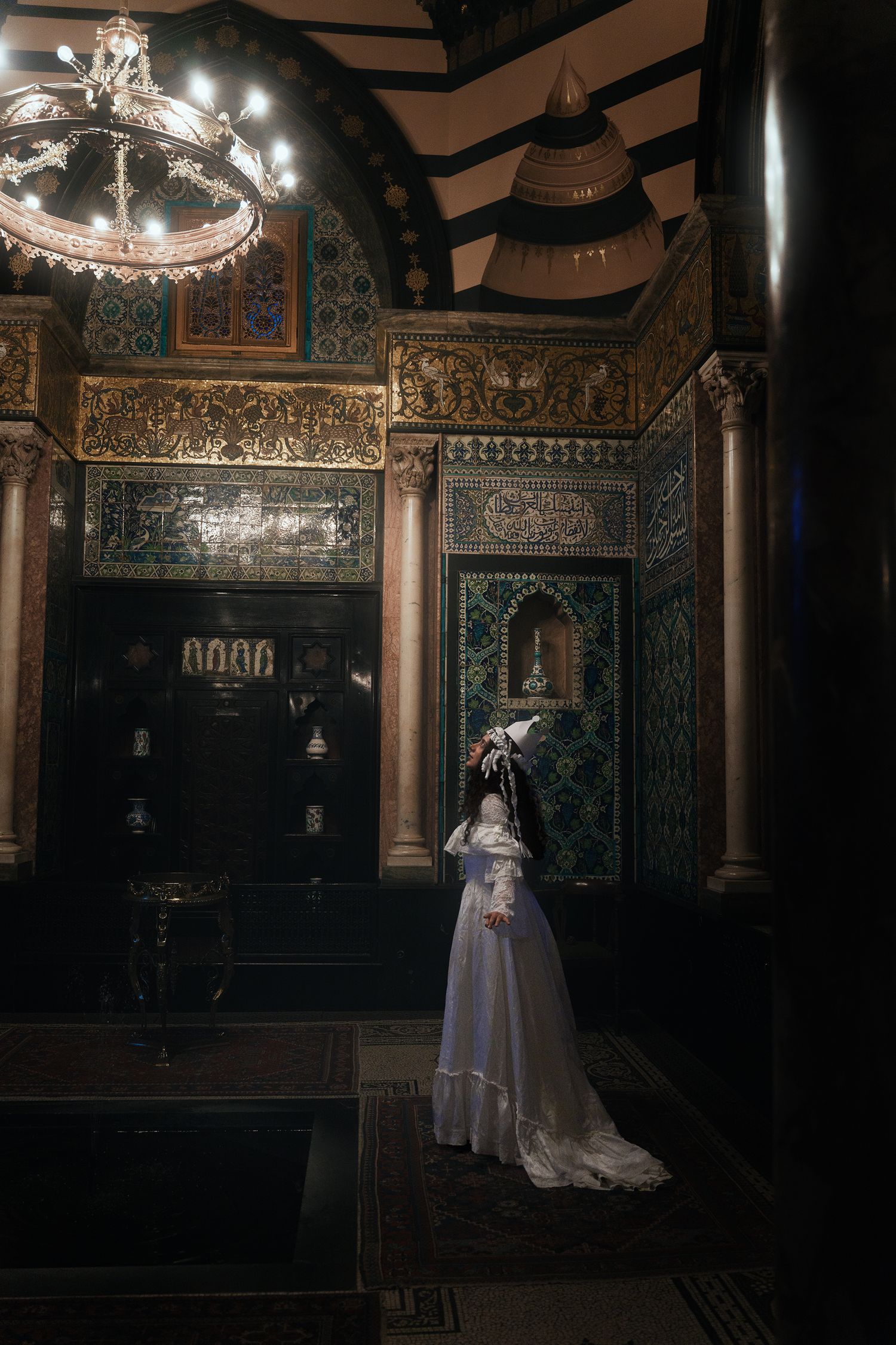 Woman in white gown stands before elaborate Islamic tilework and chandelier in richly decorated historic palatial interior.