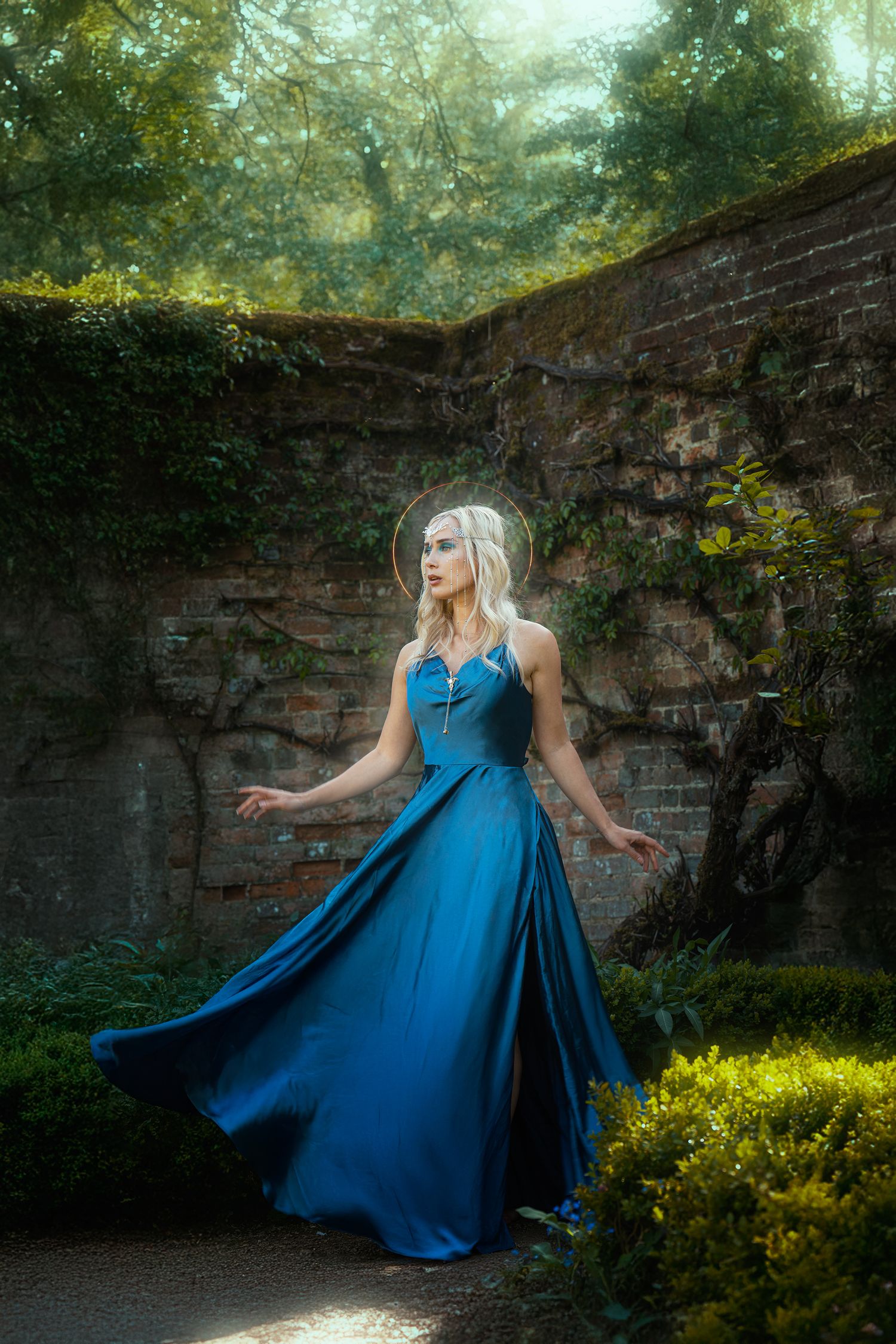 A blonde woman in a flowing teal blue gown poses gracefully in a sunlit ivy-covered brick garden.