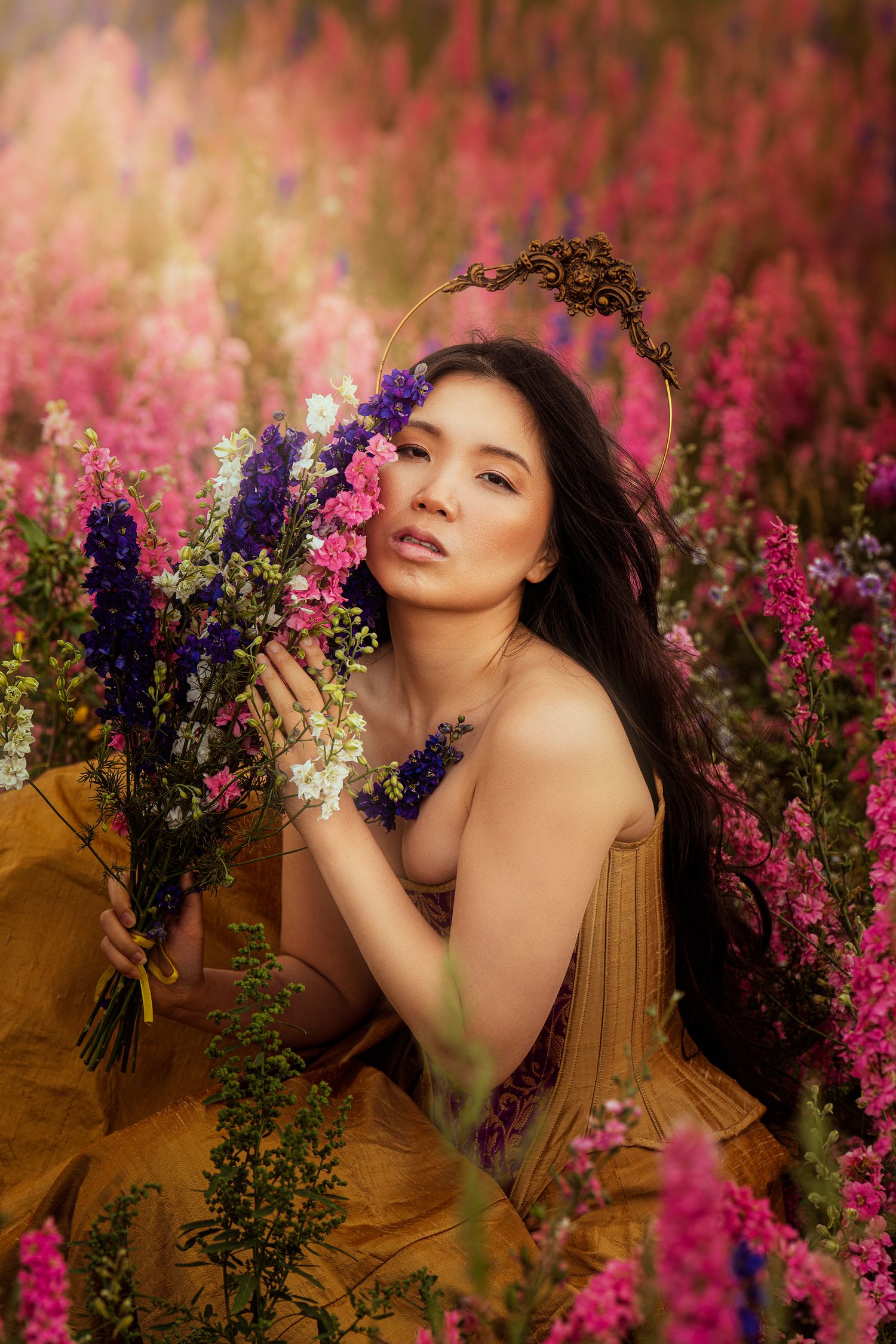 Woman in golden dress wearing ornate crown holds colorful wildflower bouquet in a pink flower field.