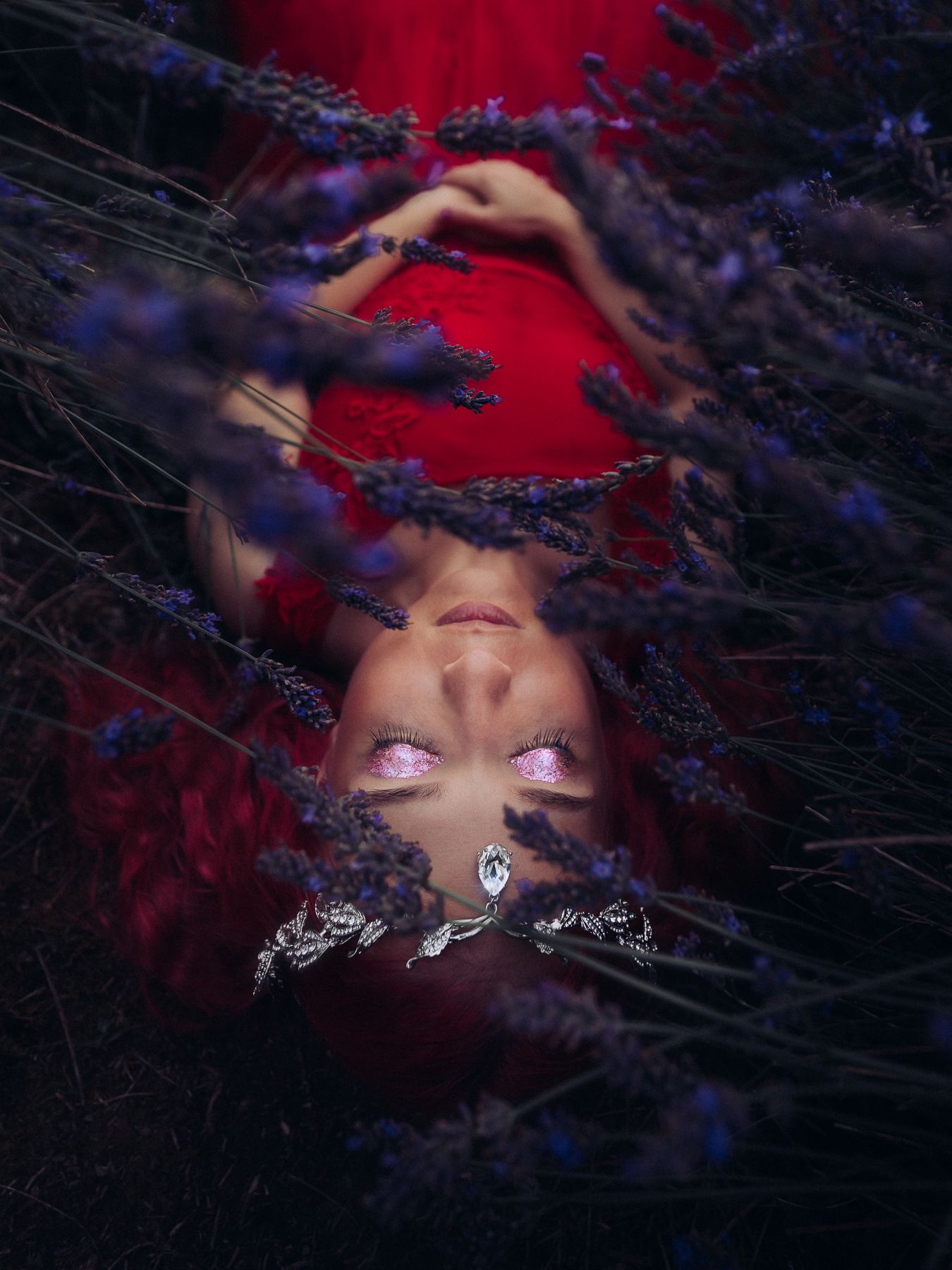 Woman with glowing eyes lying among dark purple lavender flowers wearing red dress and silver crown.