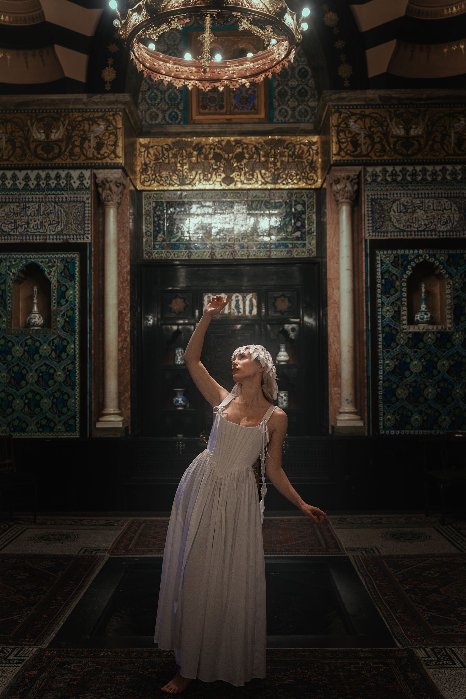 Woman in white slip dress reaches upward beneath glowing chandelier in opulent room with Islamic tilework and golden decor.