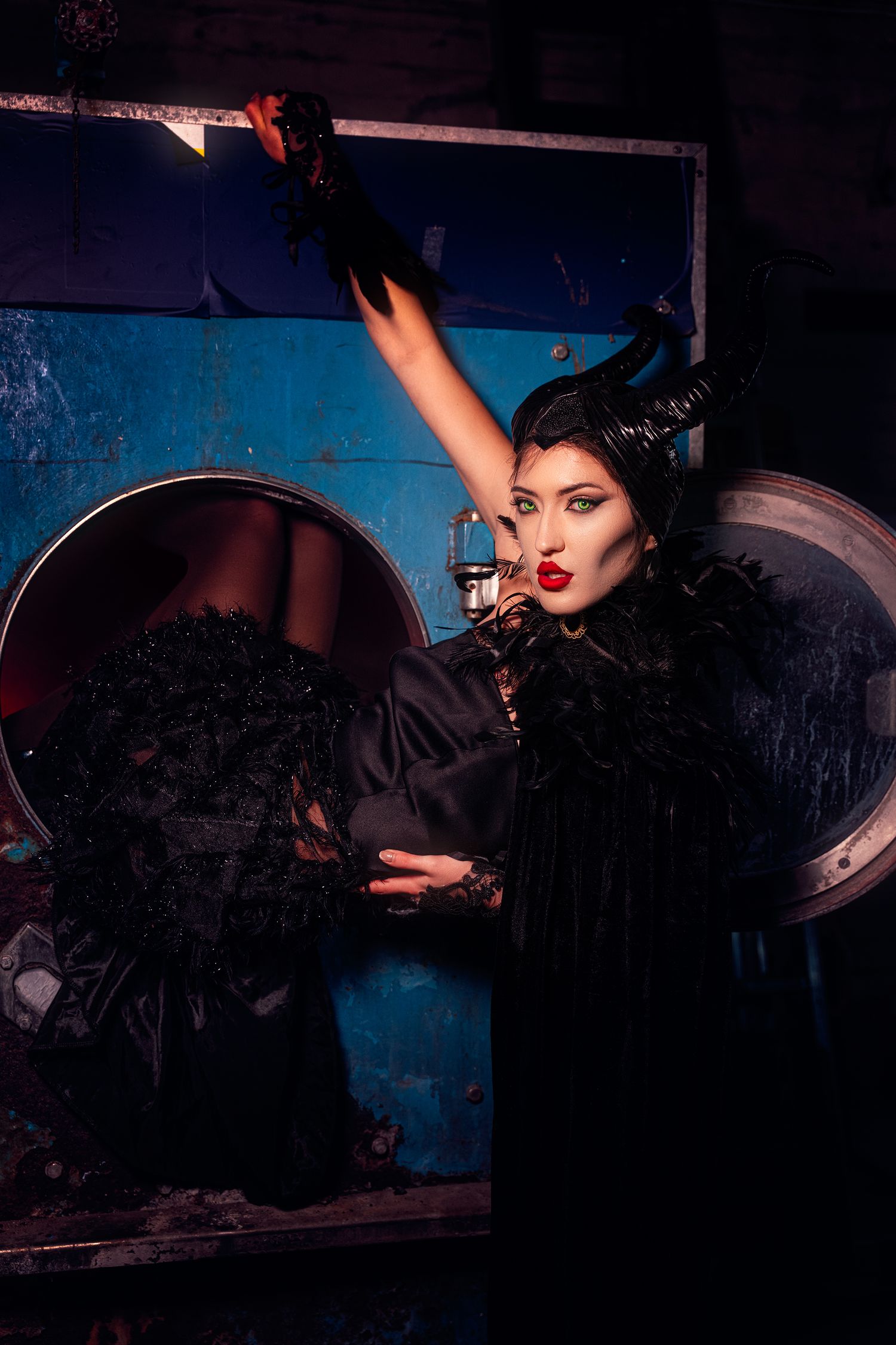 A woman in a black Maleficent costume with large horns raises her arm dramatically in a dark industrial setting.