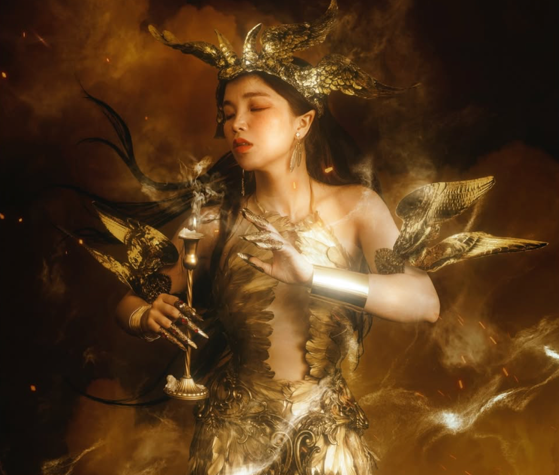 Woman in elaborate golden fantasy costume with horned headdress holding candle in dramatic golden light.