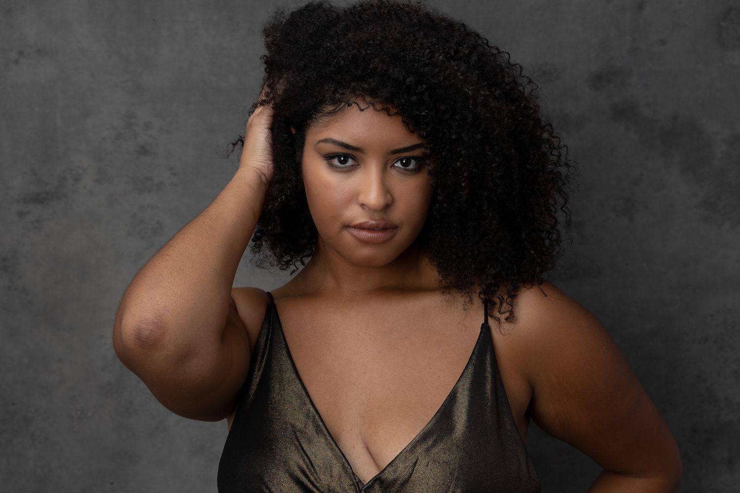 Studio portrait against gray background shows person with natural curly dark hair wearing metallic bronze top.