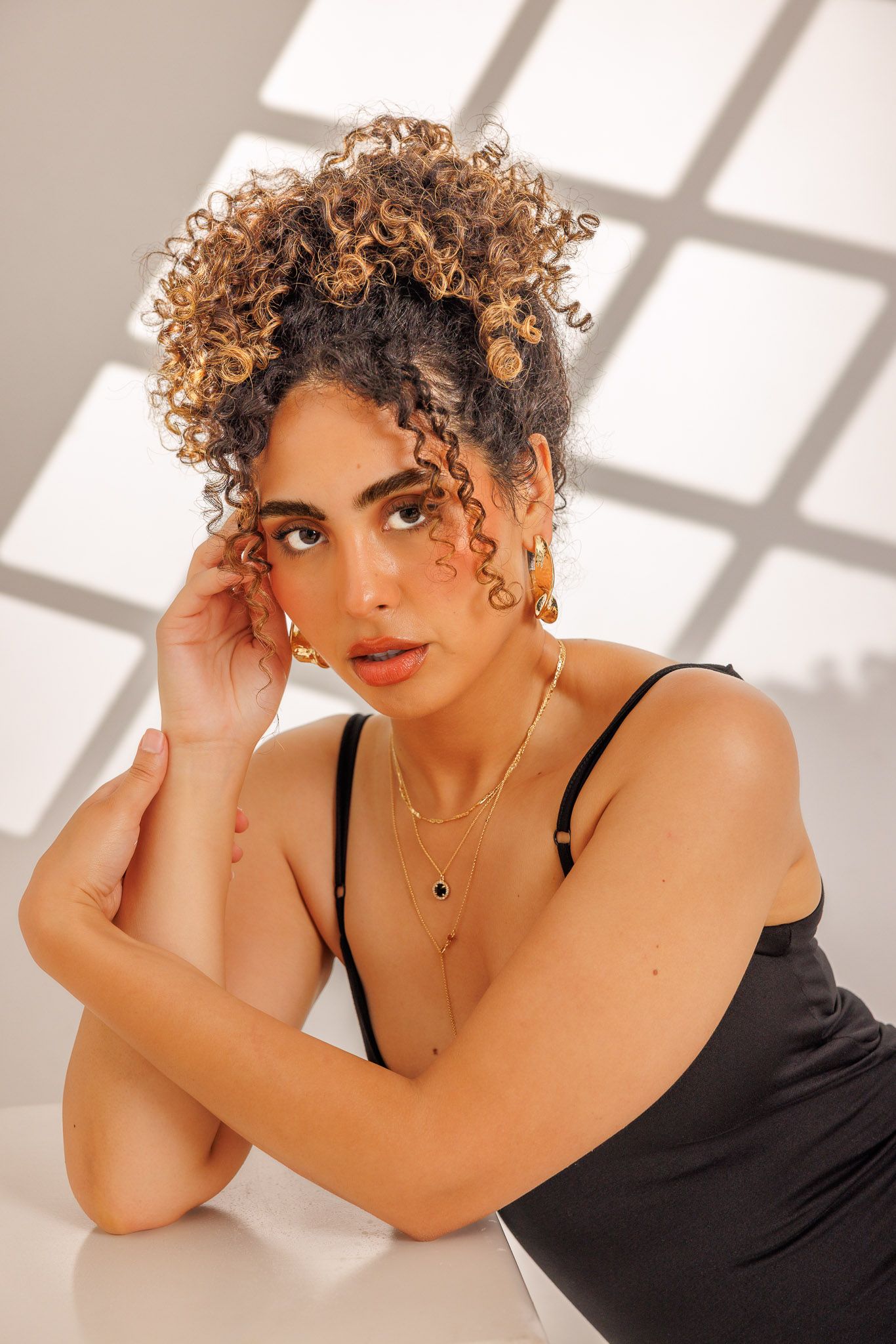 Portrait of a model with curly honey-blonde hair wearing a black tank top against a white background with window shadows.