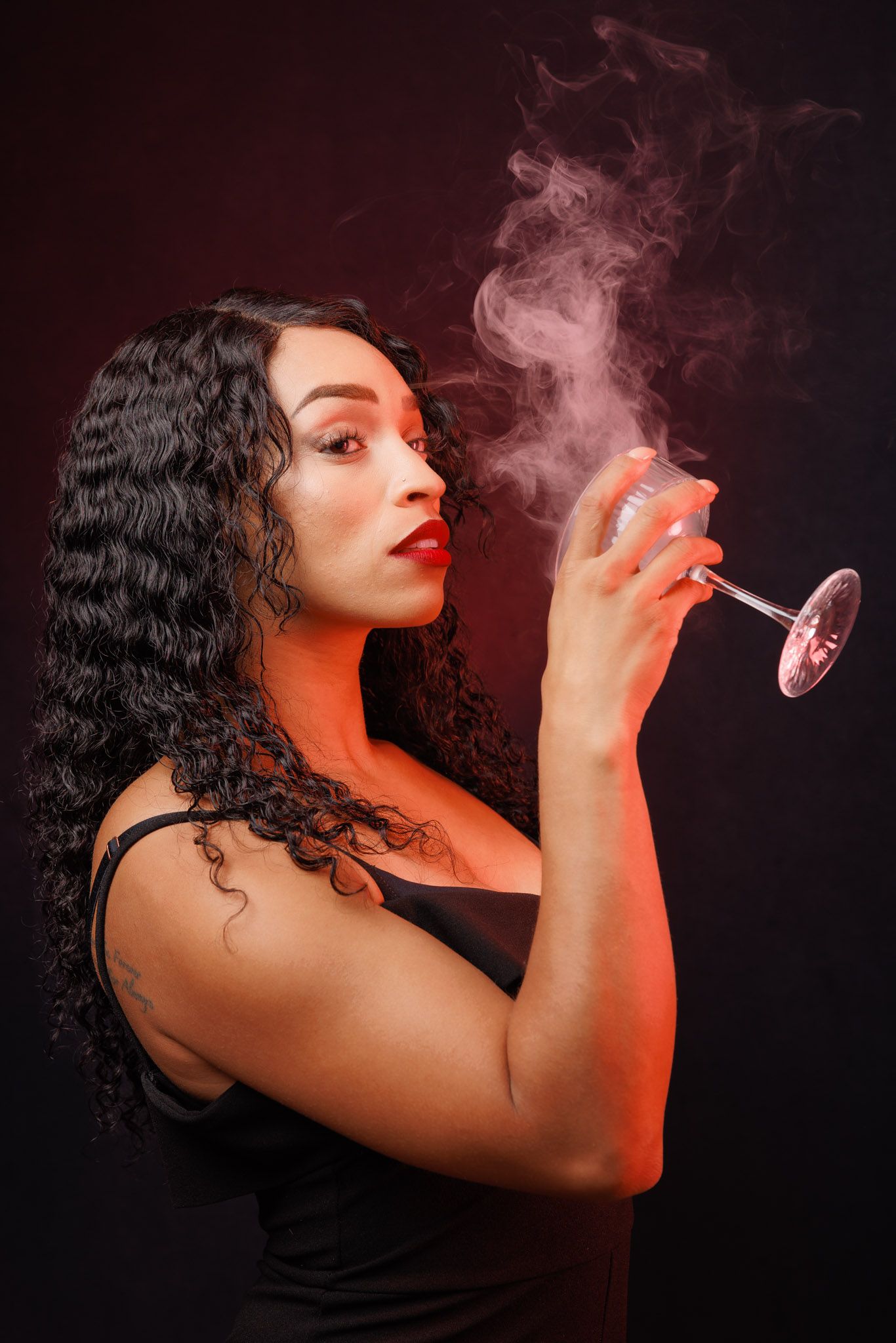 Artistic portrait with red lighting shows curly dark hair and black dress while smoke rises from a wine glass held elegantly.